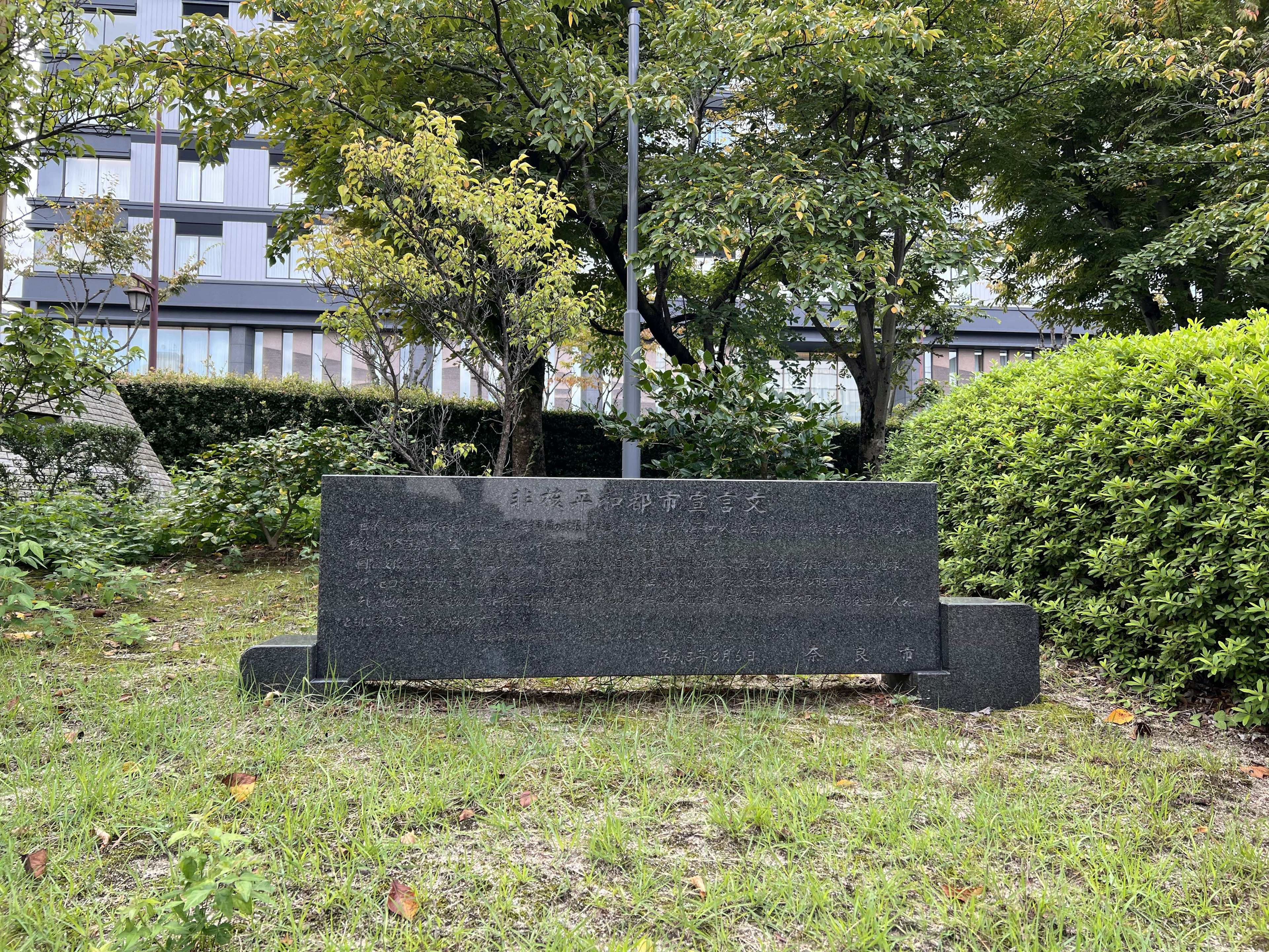 Un banco de piedra negra en un parque rodeado de césped verde y árboles con un edificio al fondo