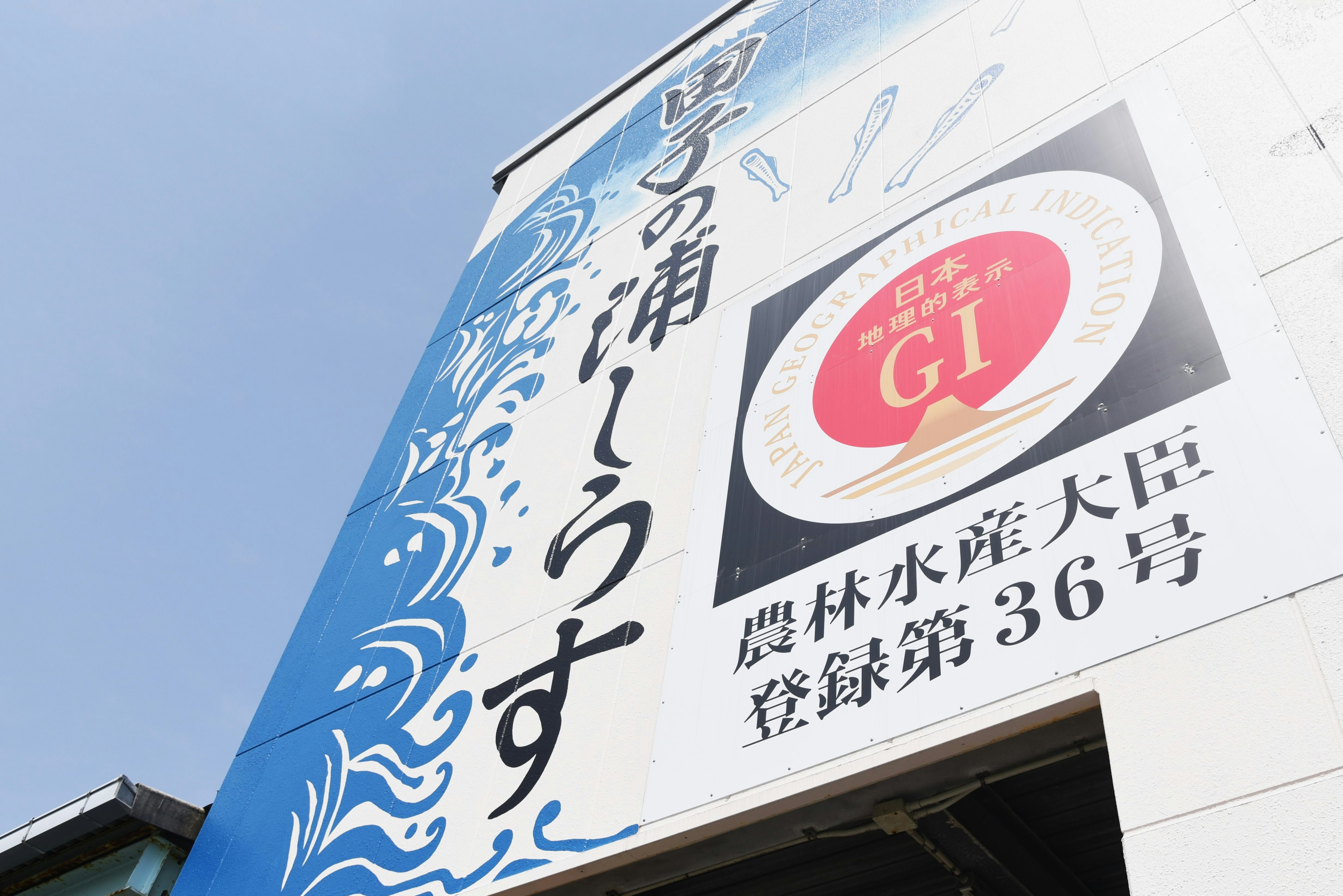 ป้ายที่มีพื้นหลังสีน้ำเงินที่มีการออกแบบคลื่นและข้อความ 'Yokkaichi no Ura' และ 'GI'