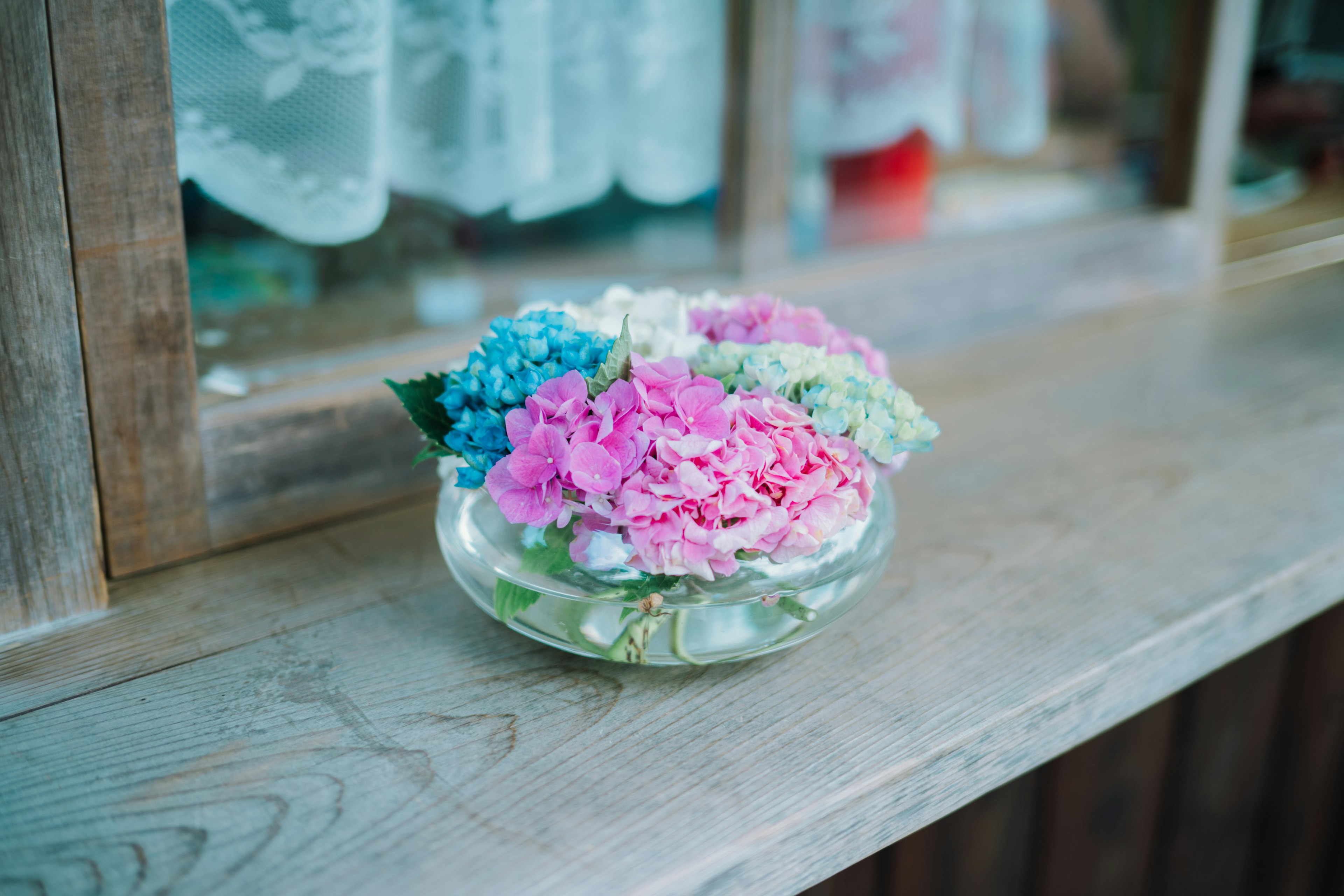 Bunte Blumen in einer klaren Schüssel auf einer Holzoberfläche neben einem Fenster arrangiert