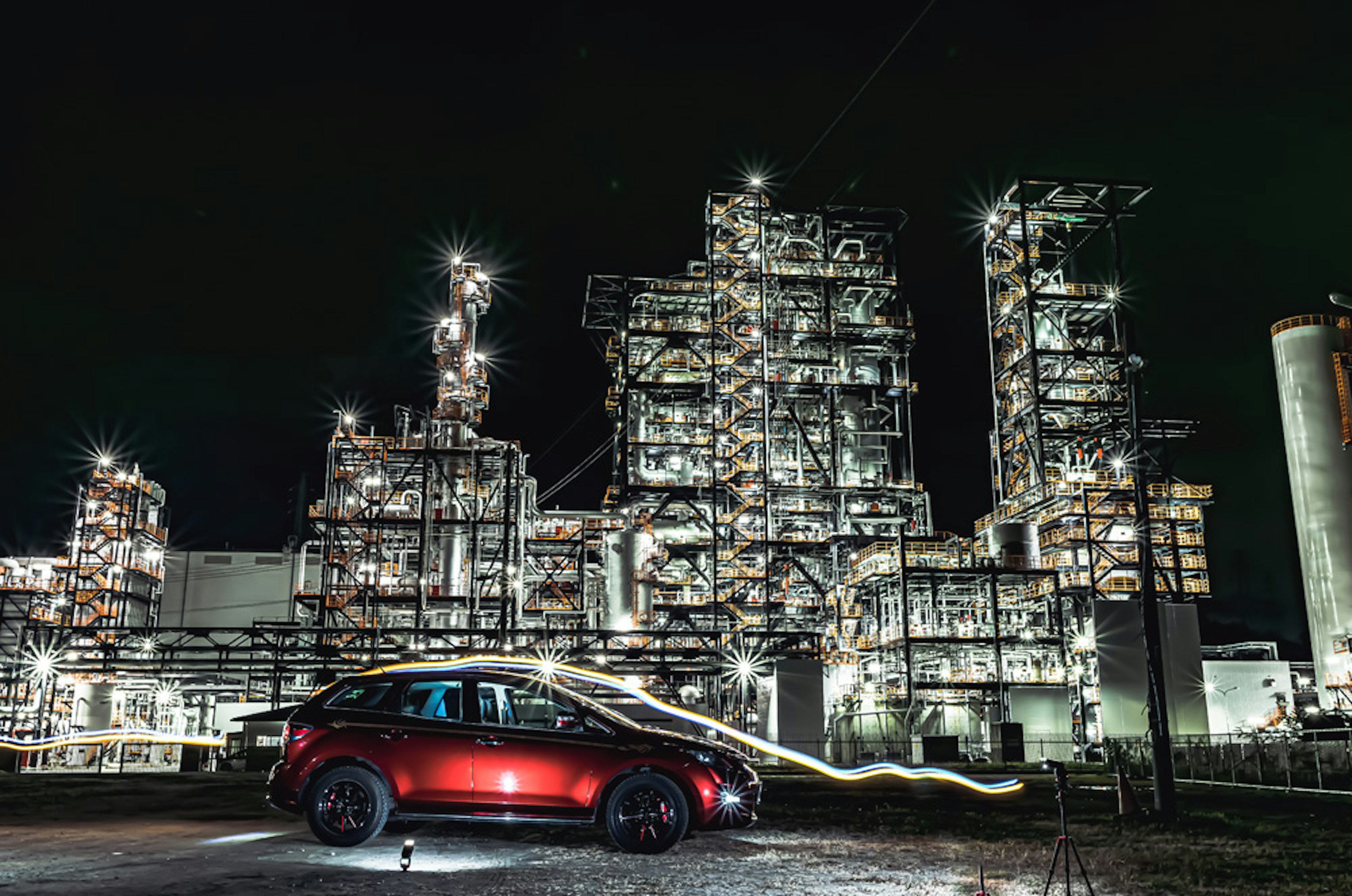 A red car parked in front of a brightly lit industrial complex at night