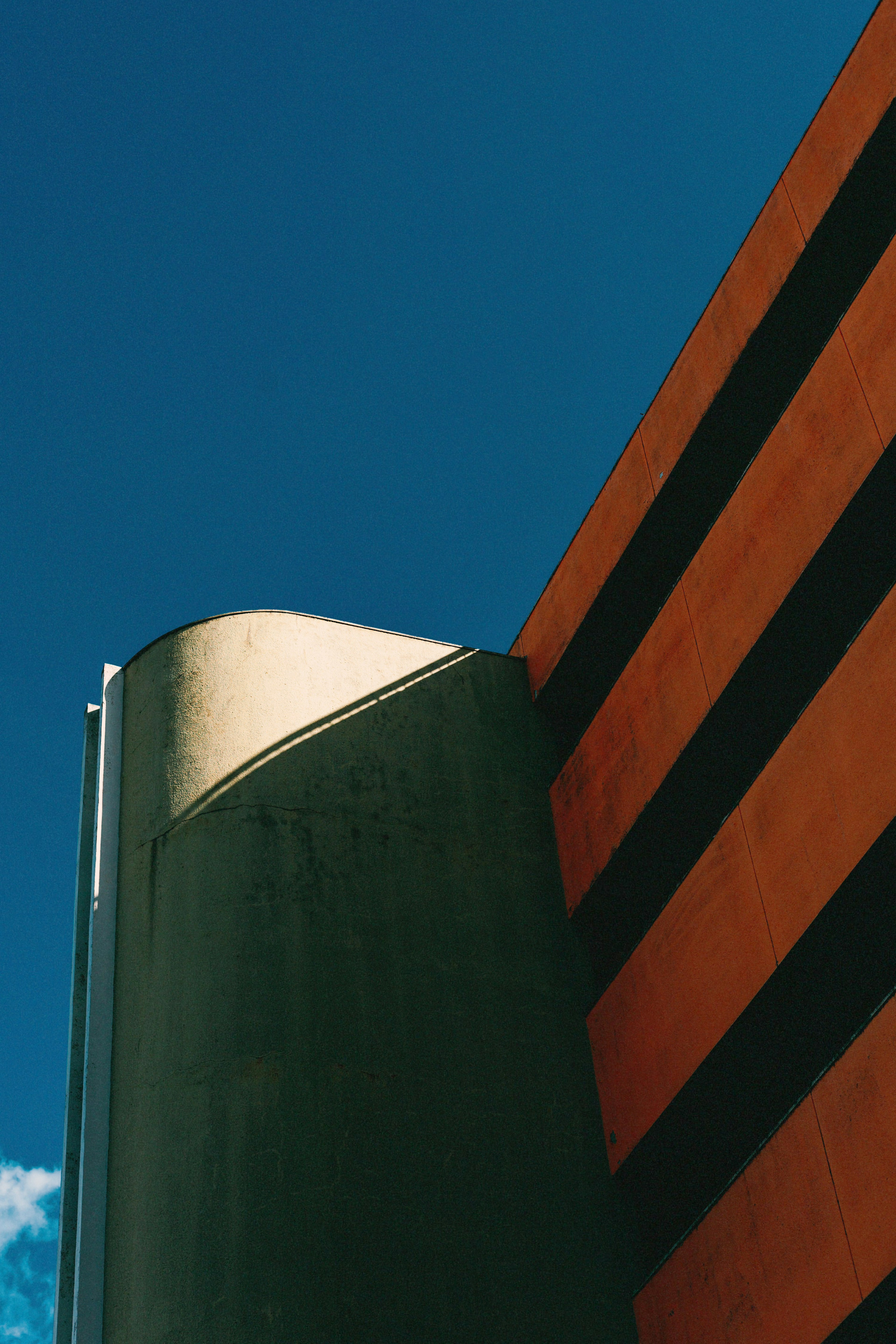 Green cylindrical structure with a curved top against a blue sky and orange striped wall