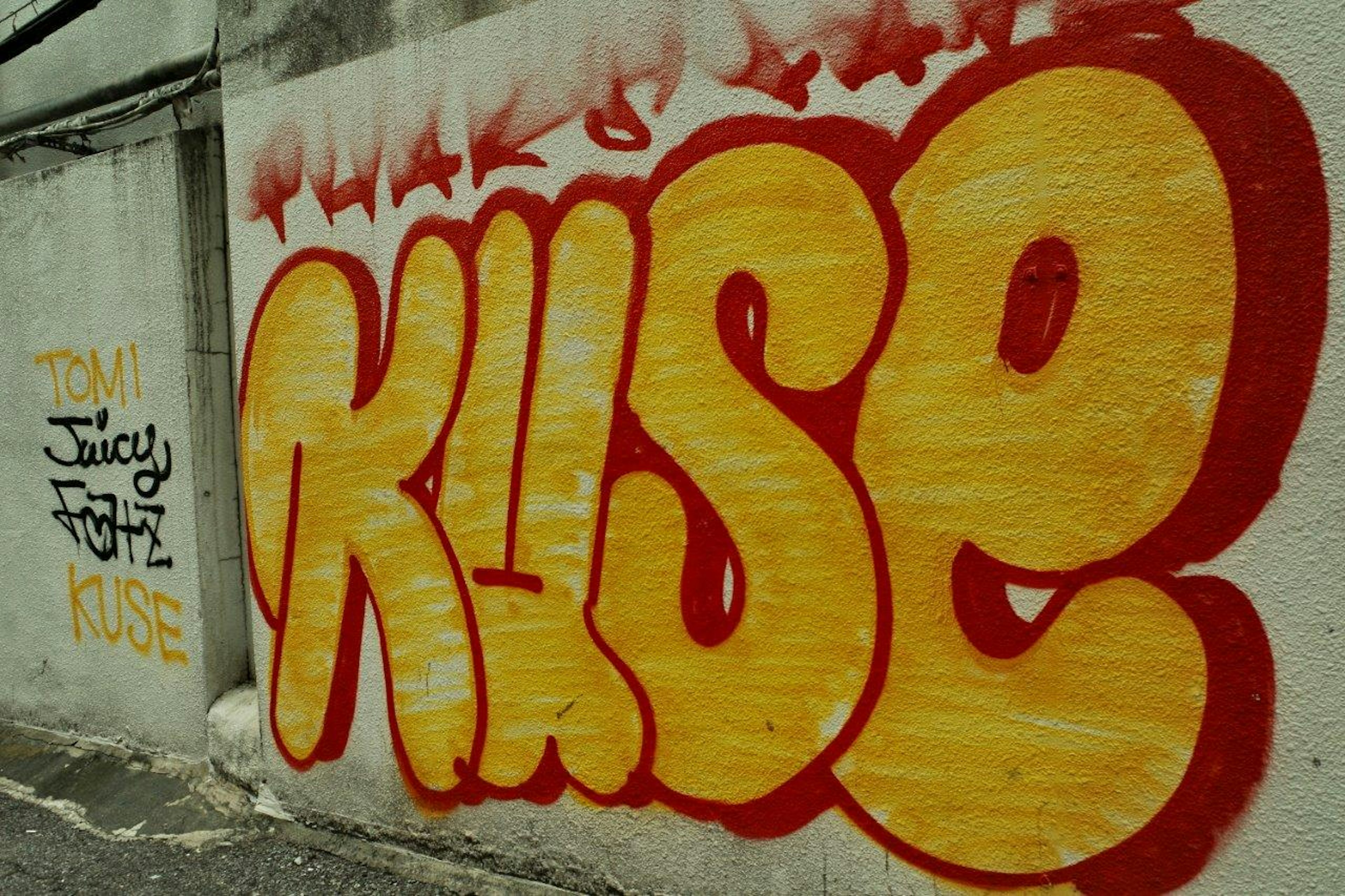 Seni grafiti besar berwarna kuning di dinding dengan kata KUSE