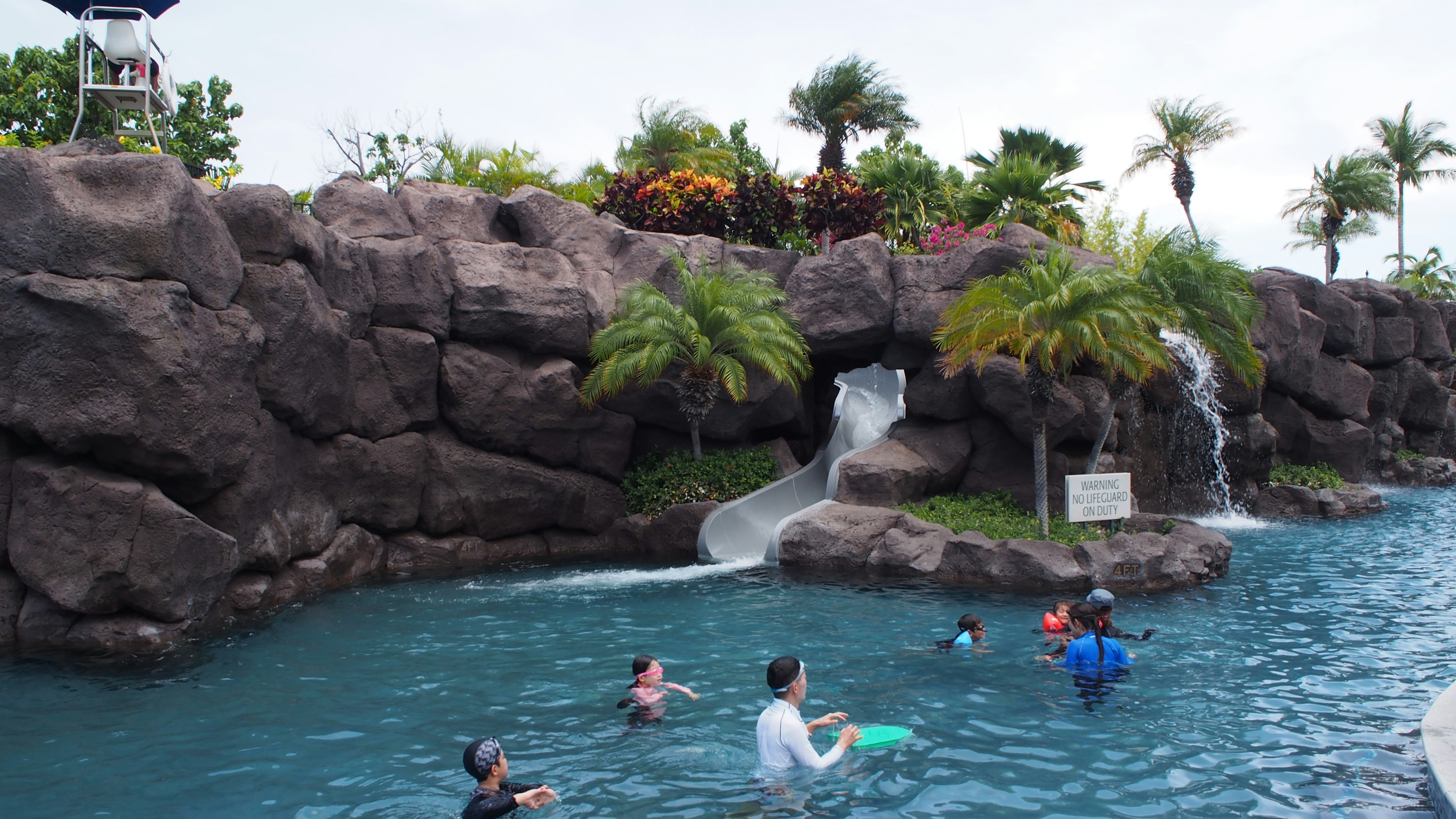 Enfants jouant dans une piscine avec un toboggan et des plantes tropicales luxuriantes