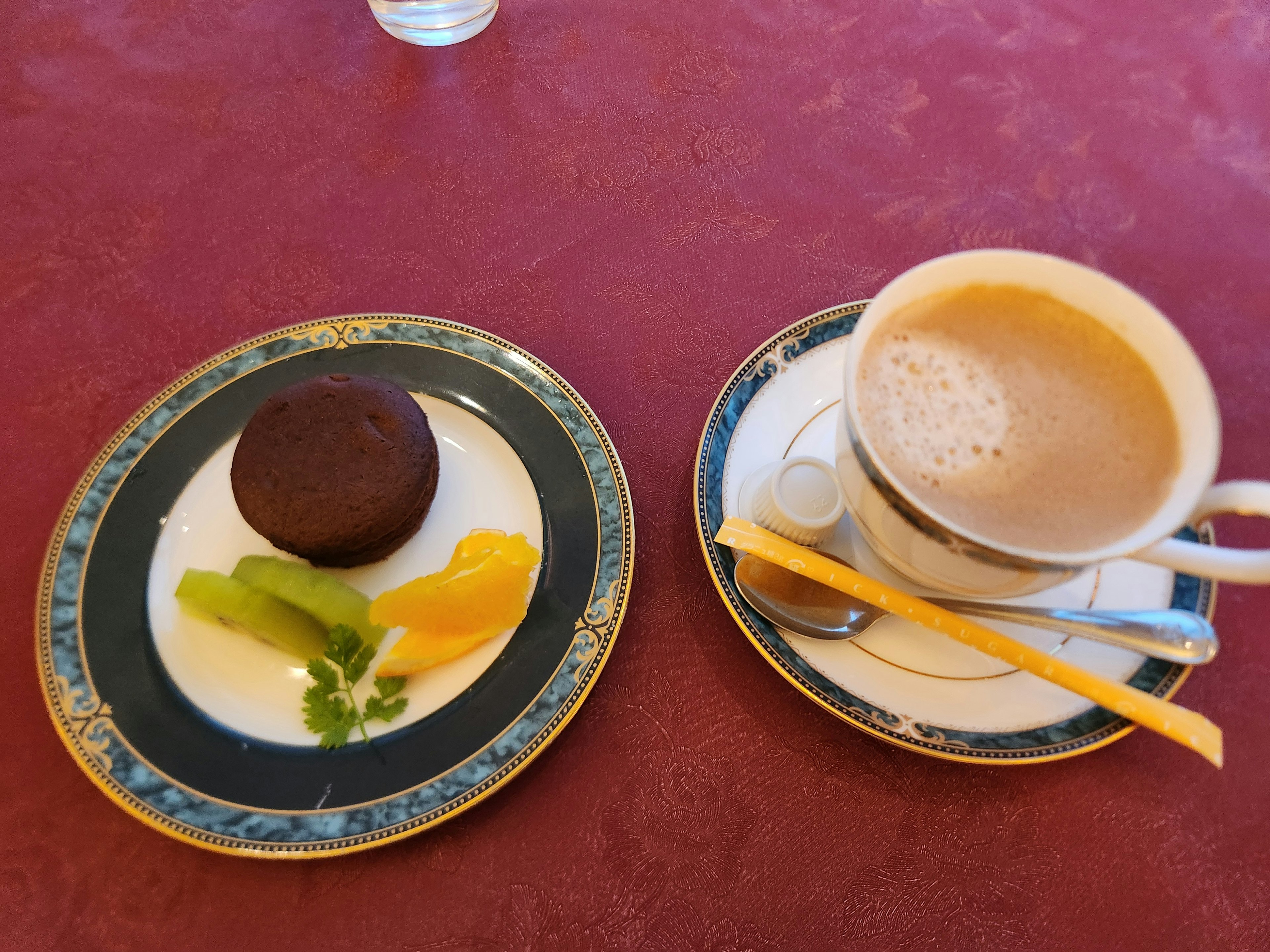 Un plato con un postre de chocolate y frutas al lado de una taza de café sobre un mantel rojo