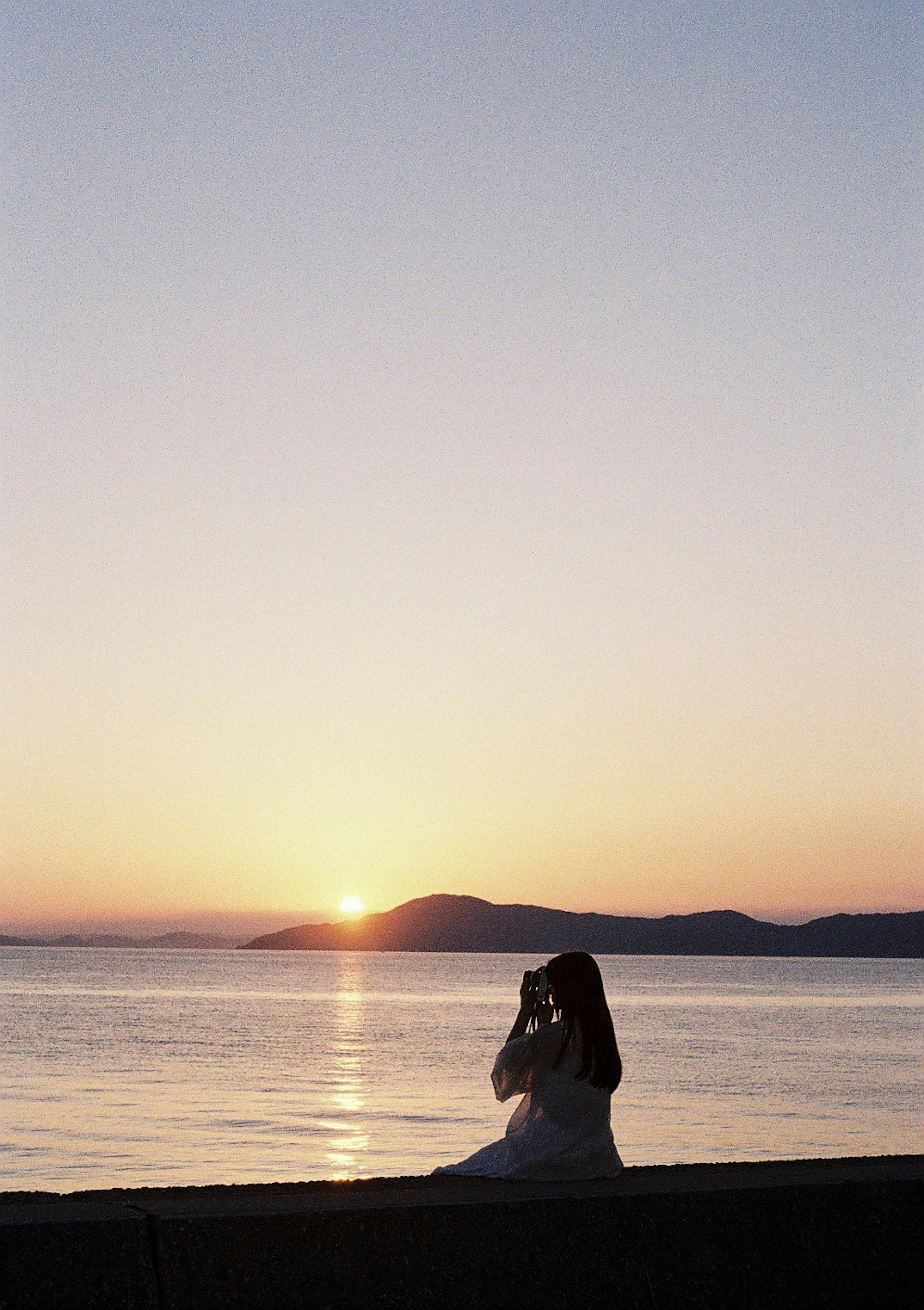 Une femme en robe blanche assise au bord de la mer contre un coucher de soleil