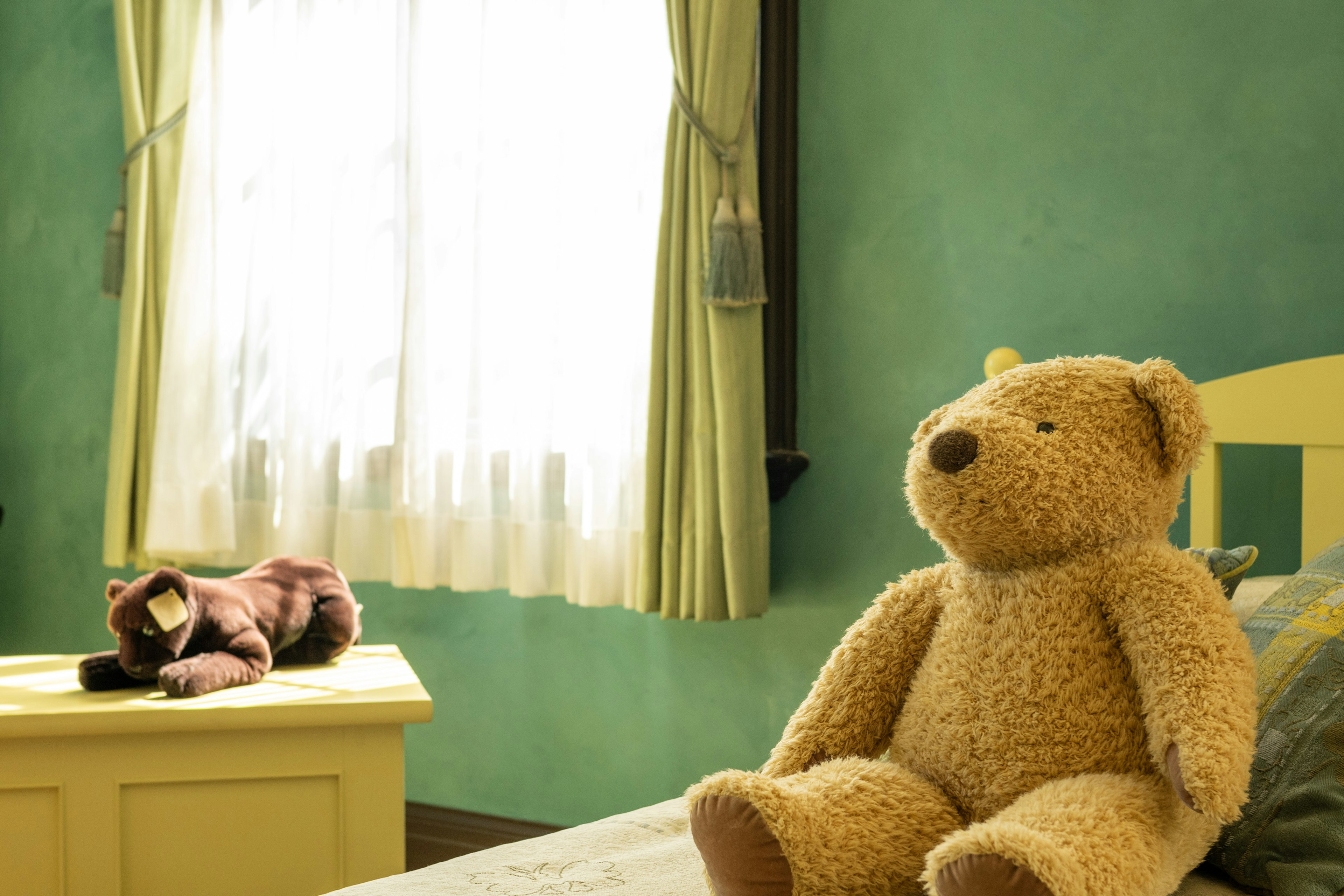 A teddy bear sitting on a bed in a child's room with green walls and curtains