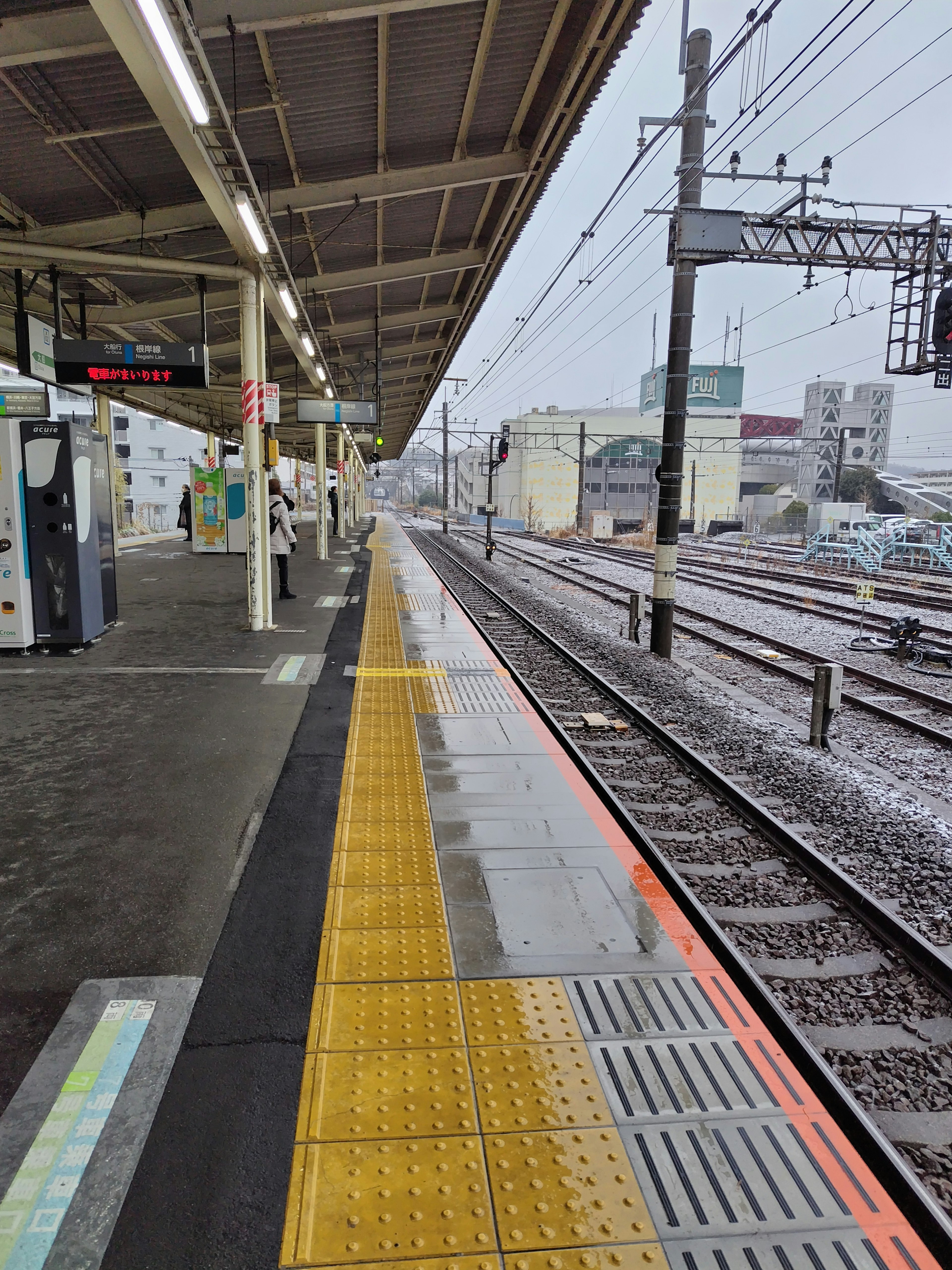 Plataforma de estación de tren con pavimento táctil amarillo y vías bajo la lluvia