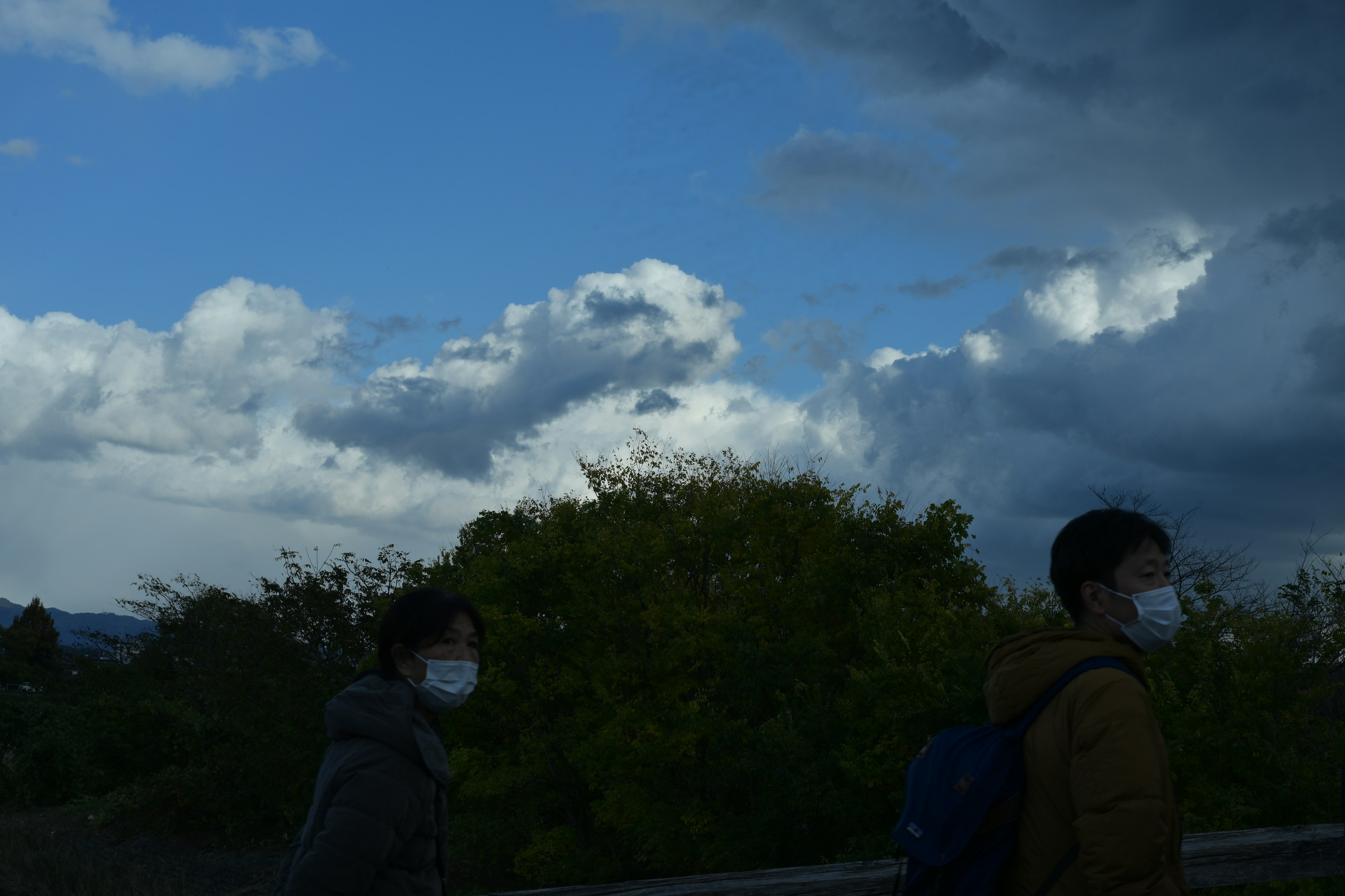 青空と暗い雲の中を歩く二人の人物がマスクを着用している風景