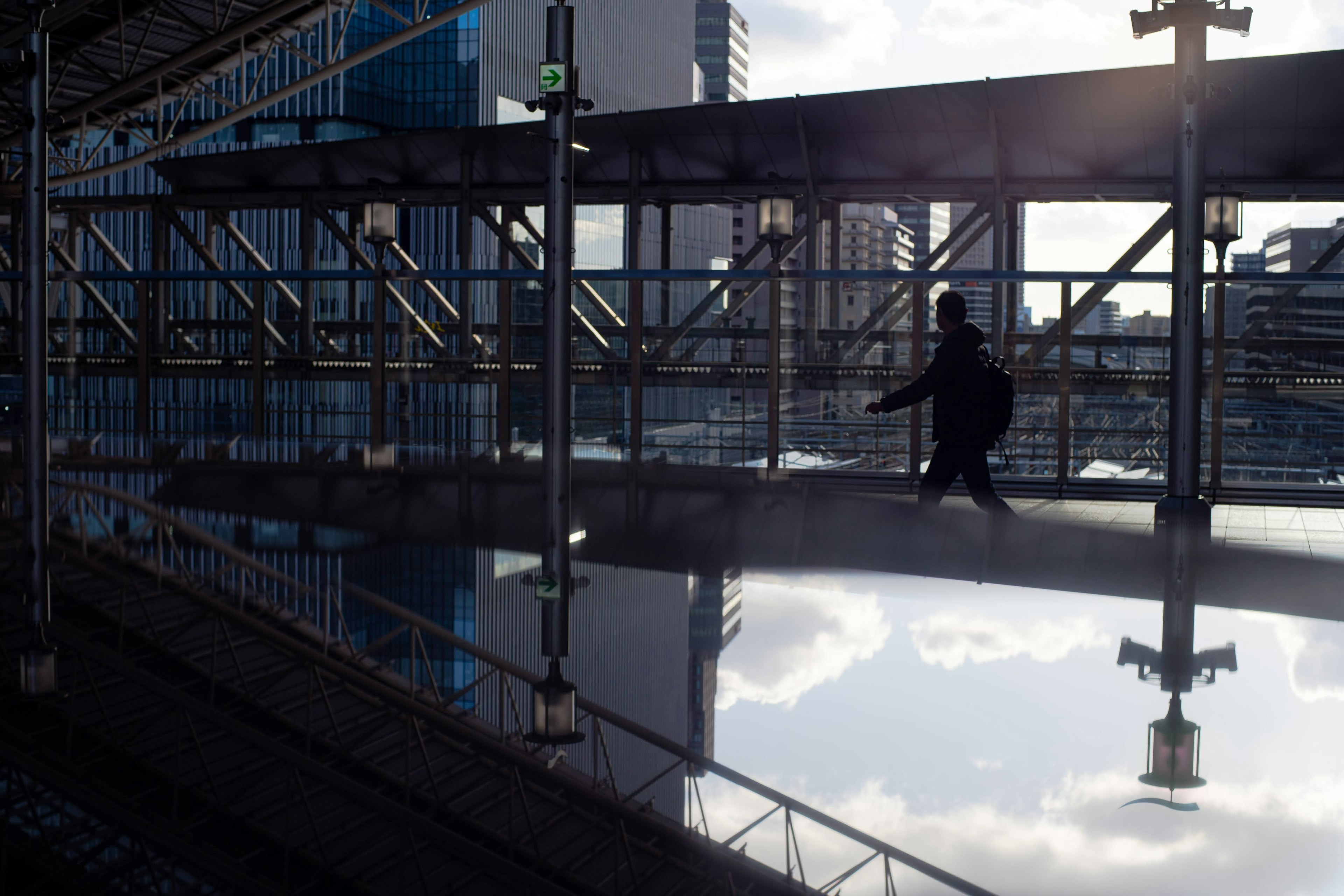 Silhouette di una persona che cammina su un ponte urbano riflessa in una superficie speculare