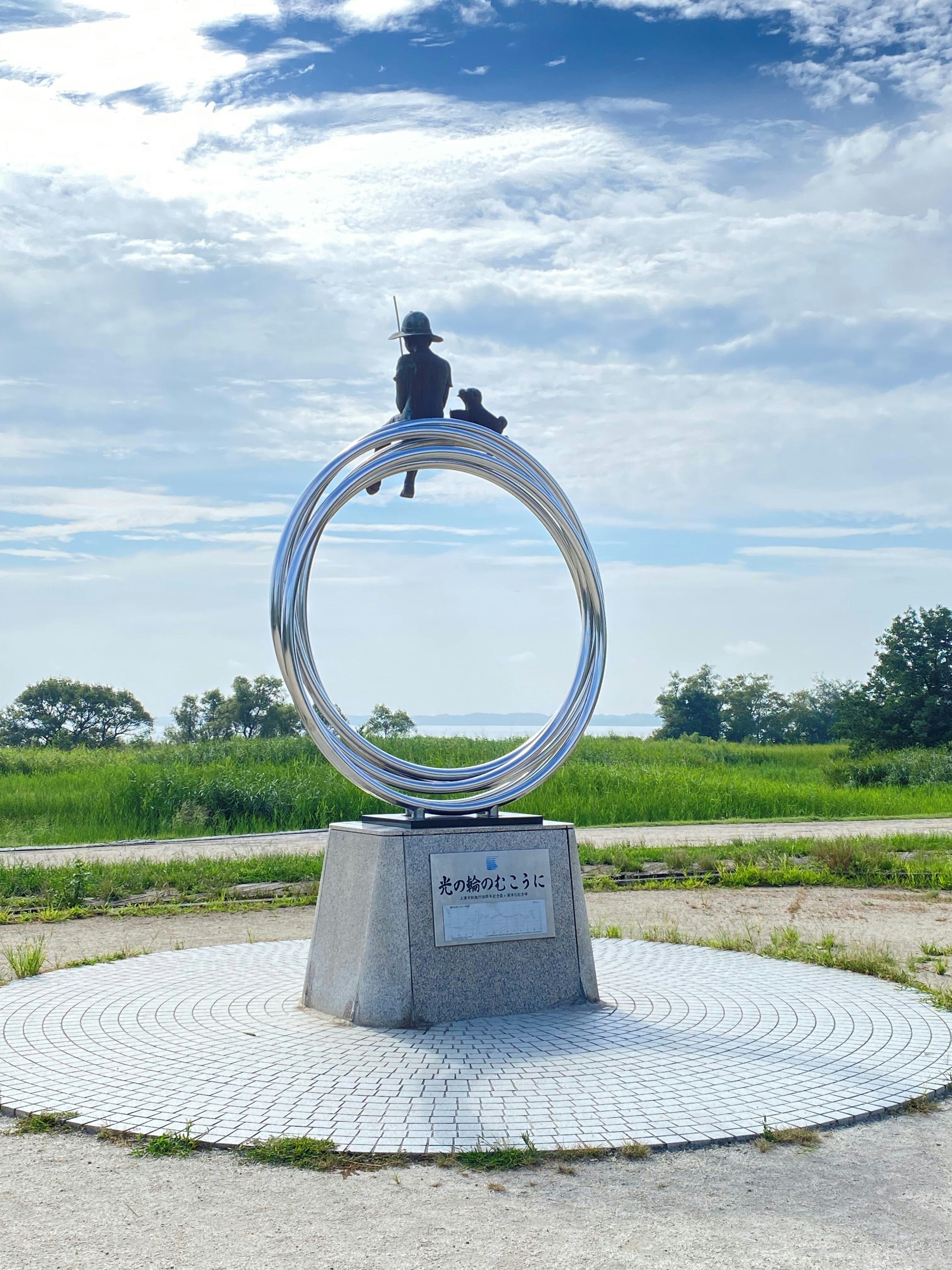 Skulptur eines Jungen und eines Hundes, die auf einem metallischen Ring unter einem schönen blauen Himmel sitzen