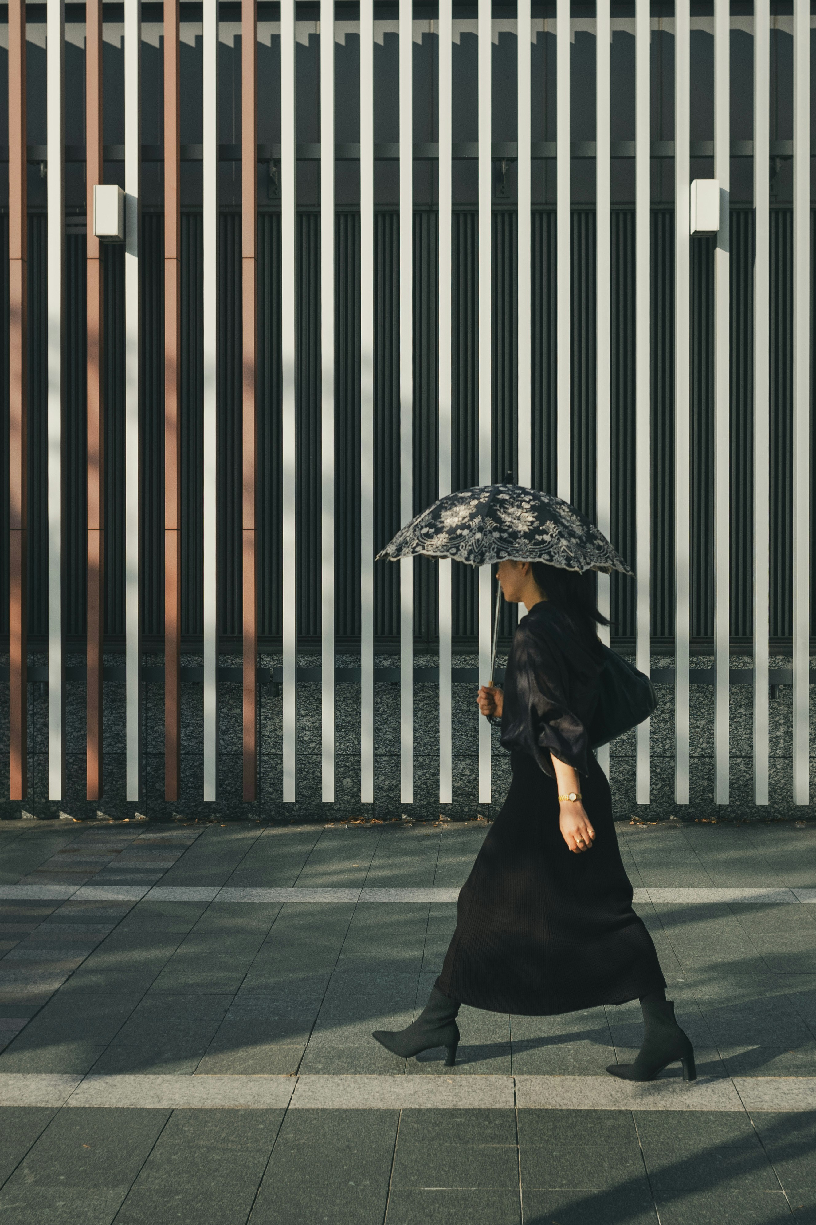 A woman in black clothing walking with an umbrella against a striped wall