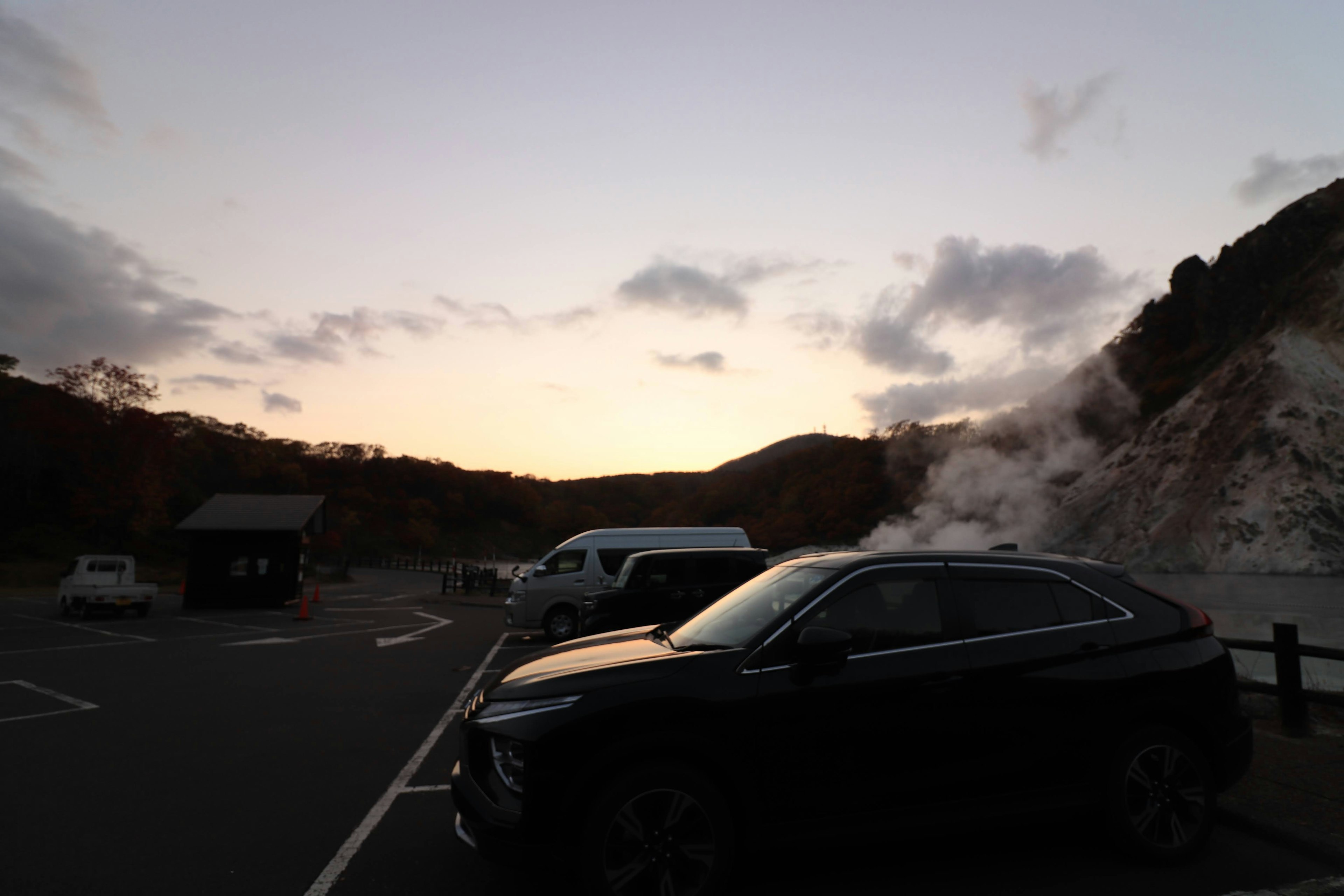 夕焼けの中、駐車場に停まった黒いSUVと温泉の蒸気が見える風景