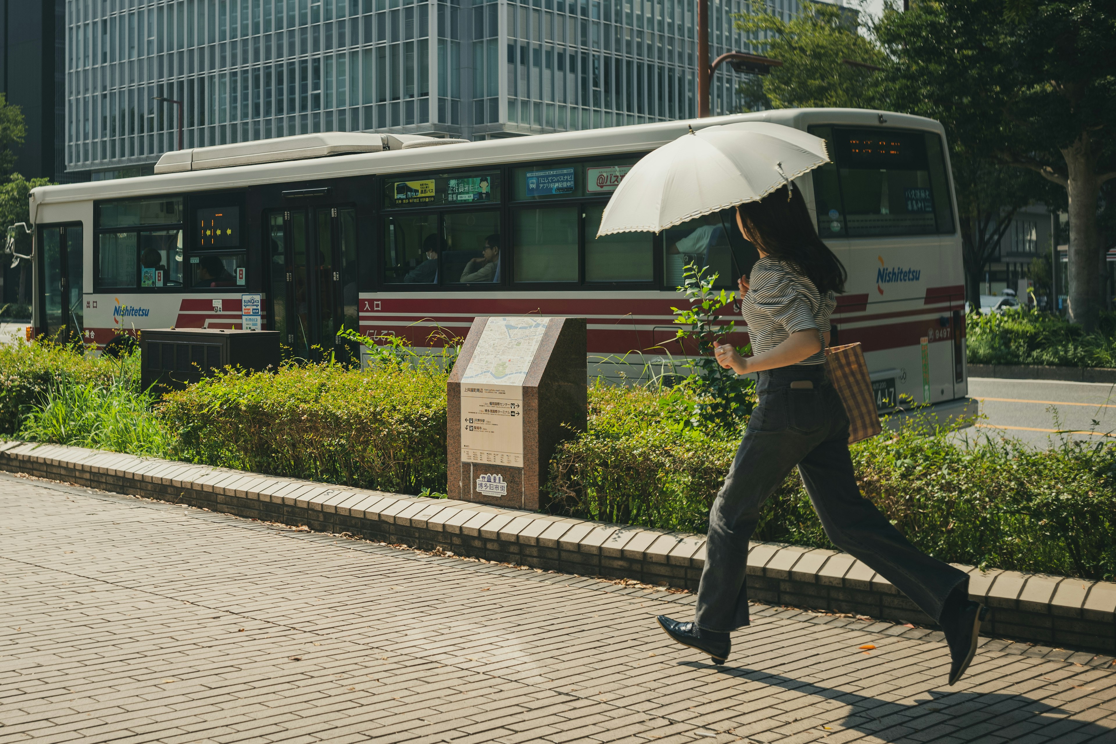 女性が傘を持って歩いている街の風景 バスが背景にあり 緑の植栽が見える