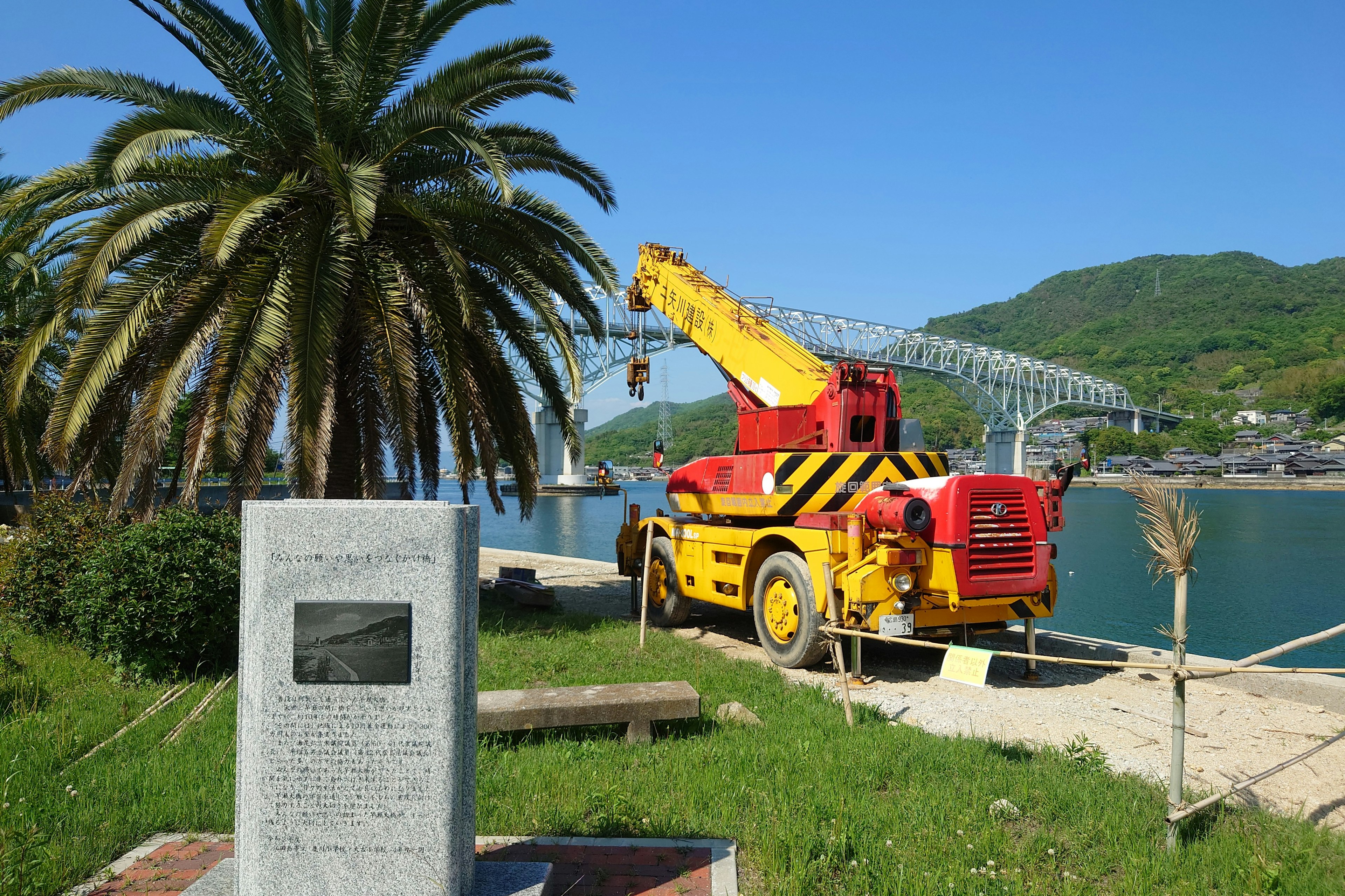 Sebuah crane merah dan kuning diparkir di tepi pantai dengan pohon palem di latar belakang