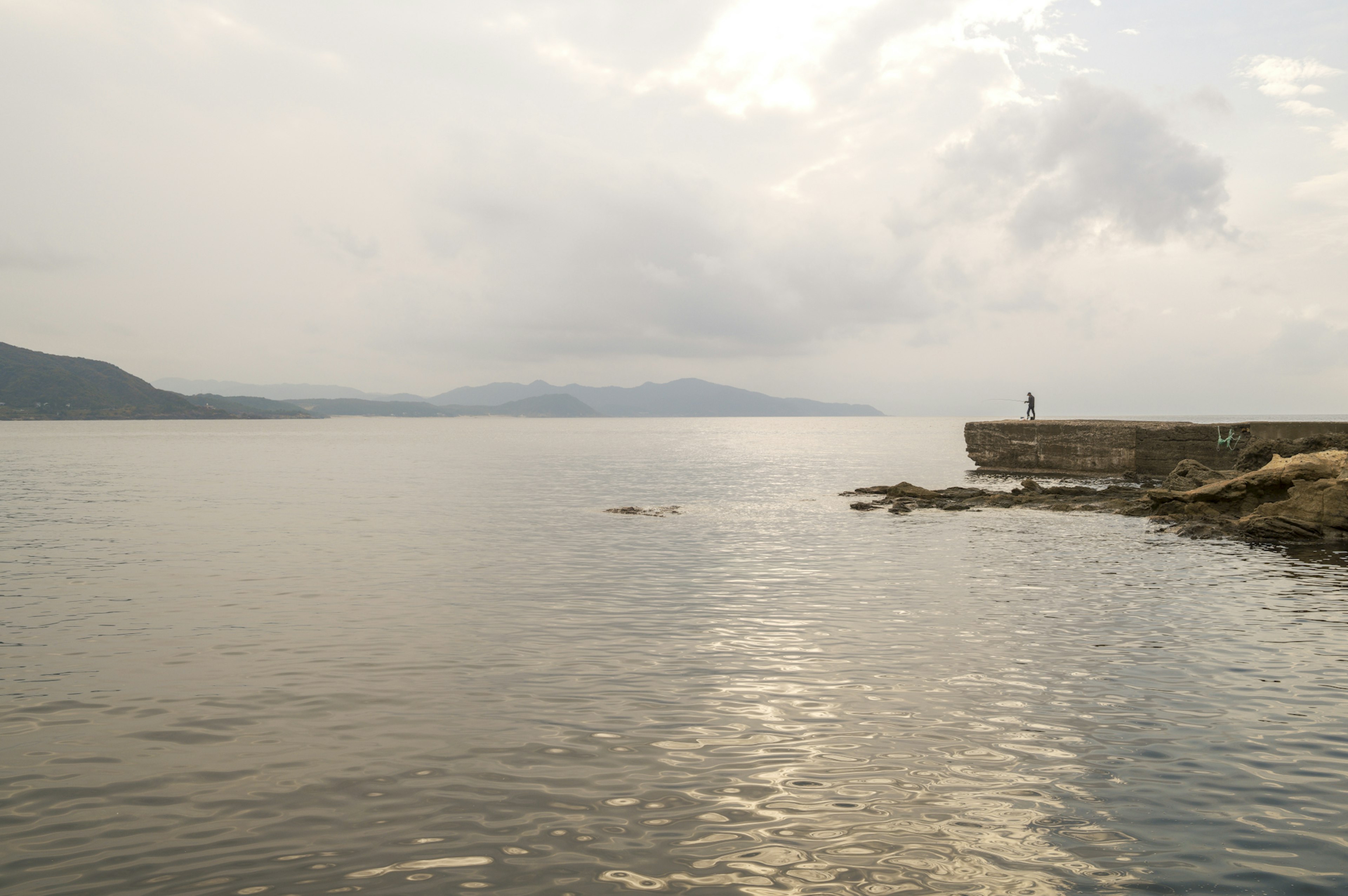 Eine einsame Figur steht auf einem felsigen Pier mit ruhigem Wasser und fernen Bergen