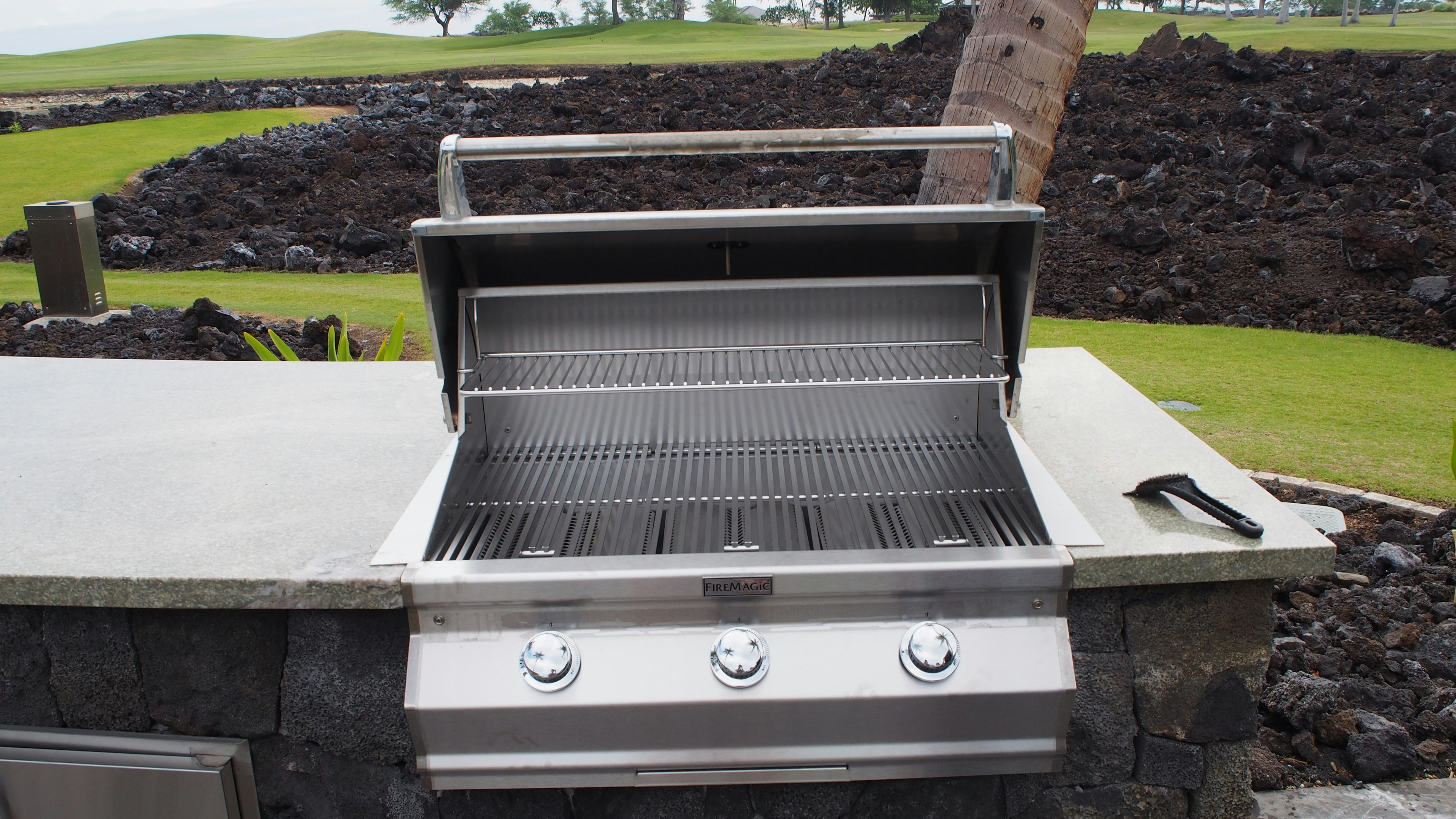 Open stainless steel grill with a stone countertop surrounded by green grass and lava rock