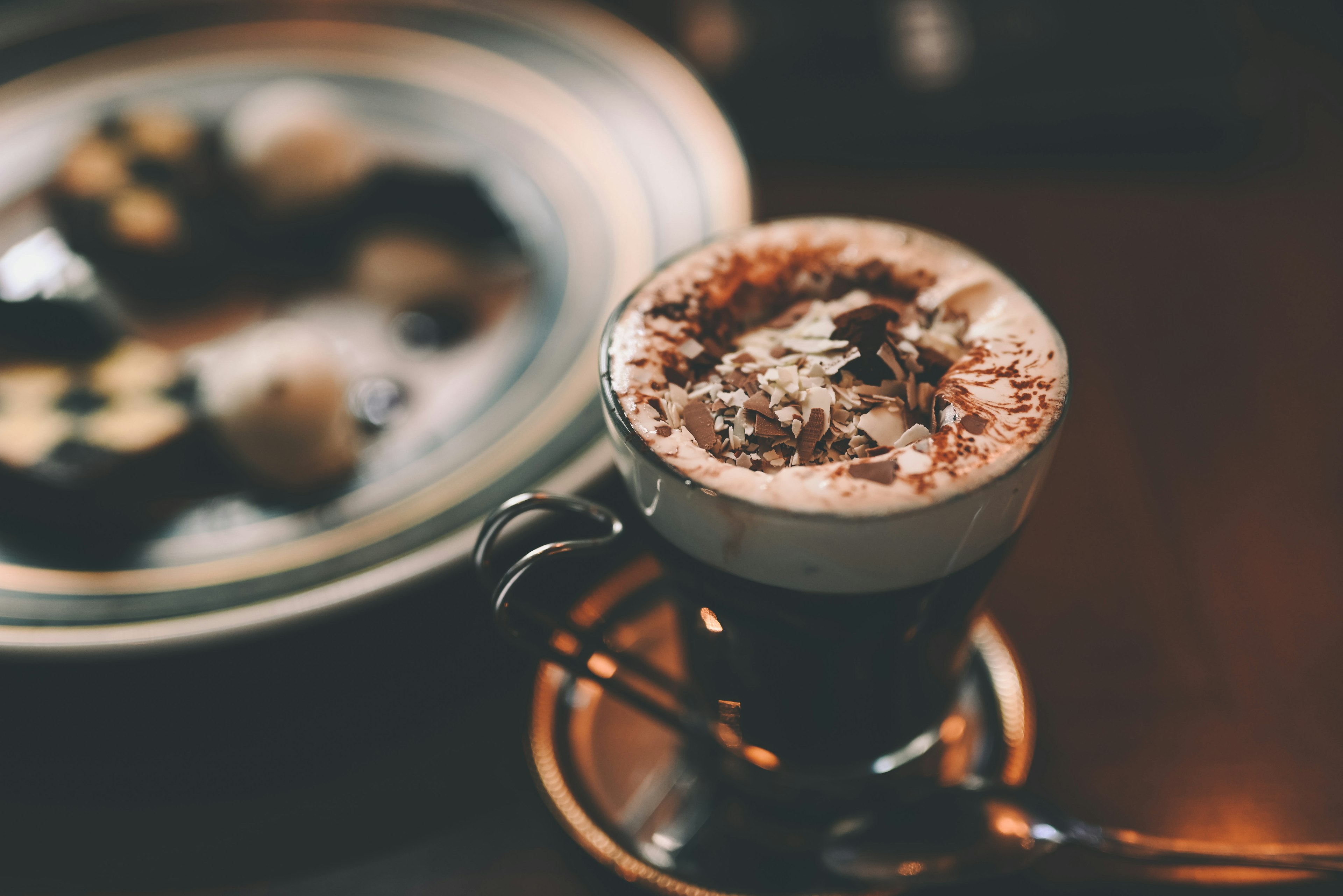 コーヒーのカップにホイップクリームとチョコレートトッピングがあり、背景にはデザートプレートがある