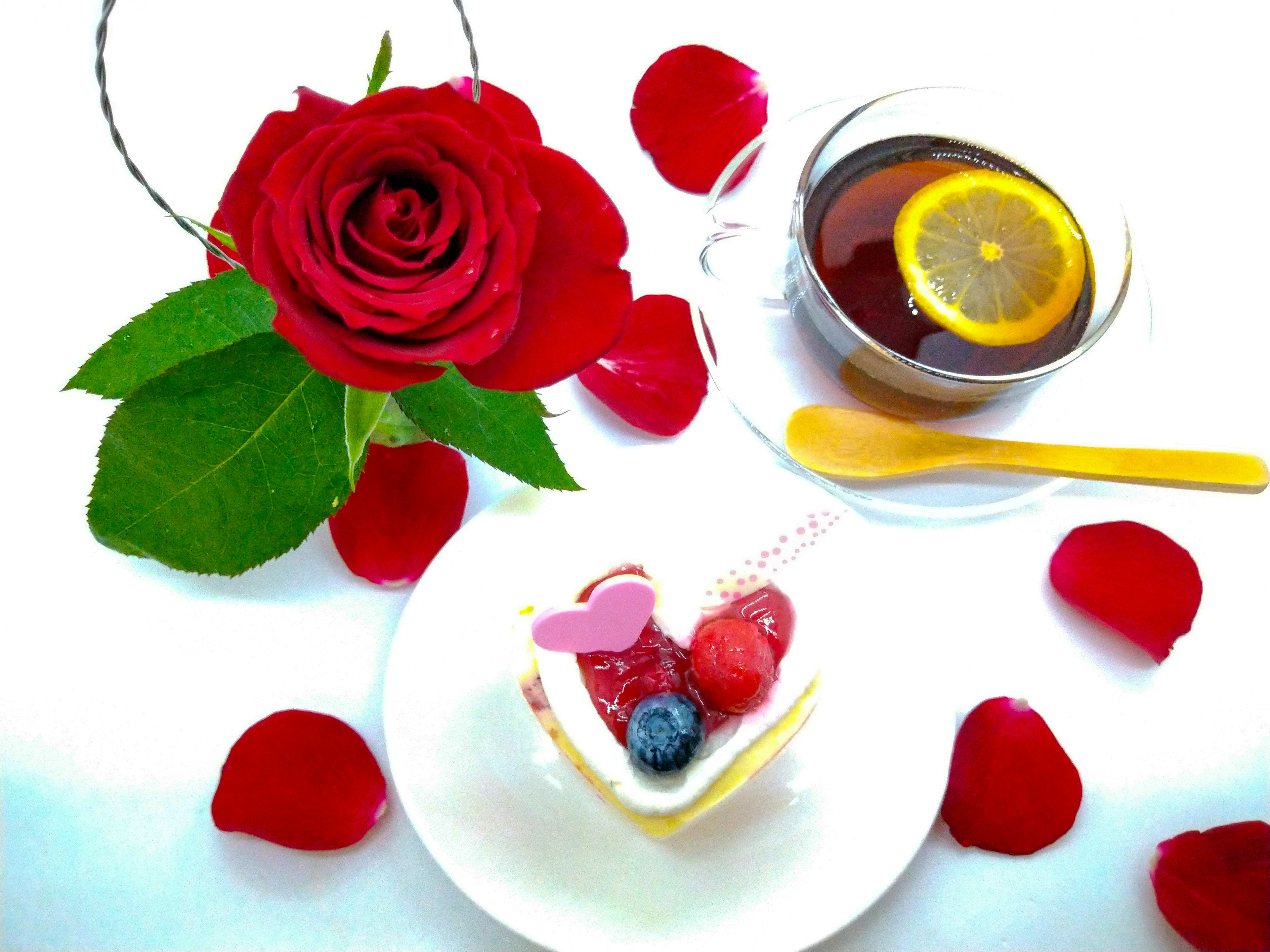 A heart-shaped cake with berries beside a red rose and petals on a white surface