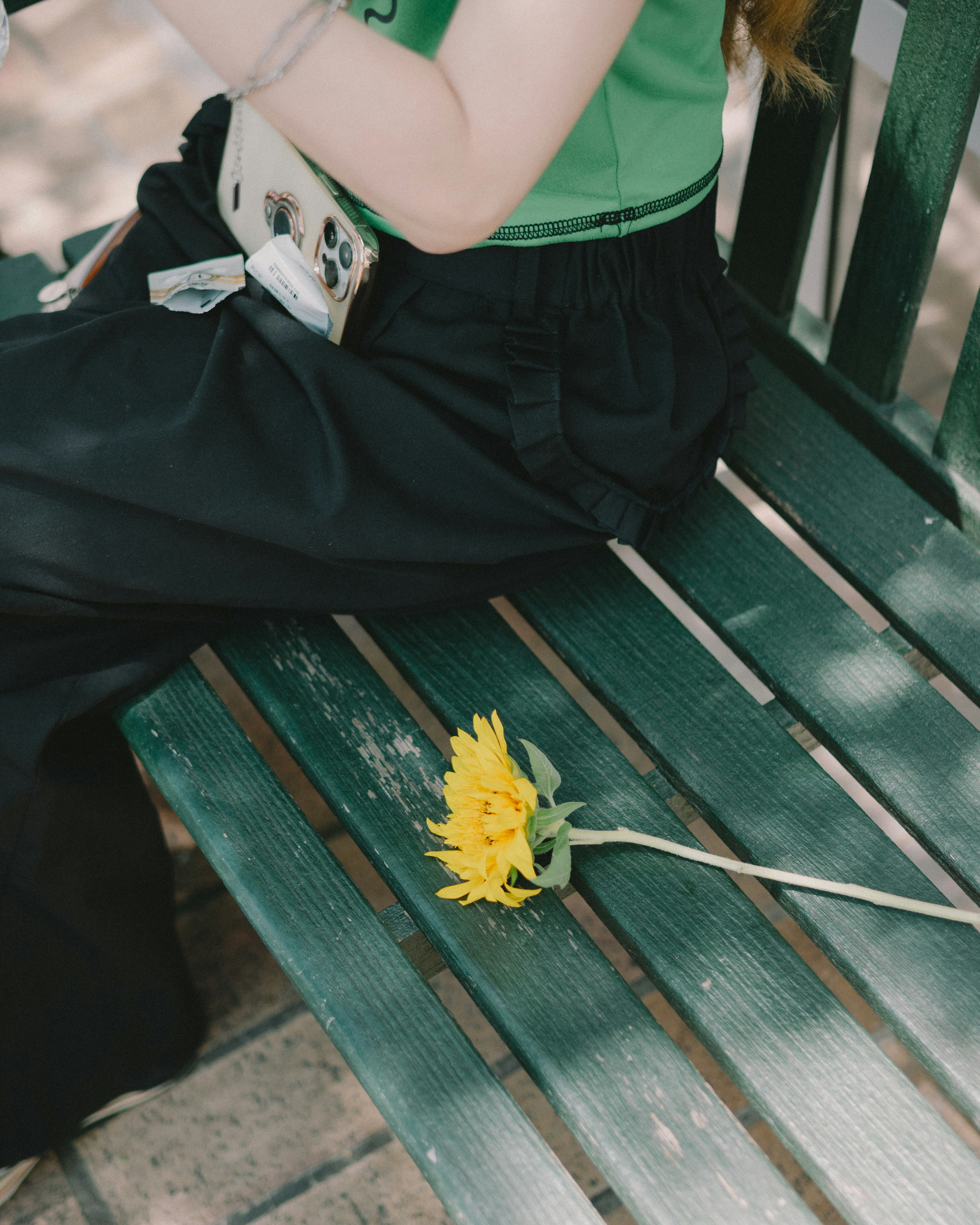 Una mujer con una blusa verde sentada en un banco usando pantalones negros con un girasol amarillo a su lado