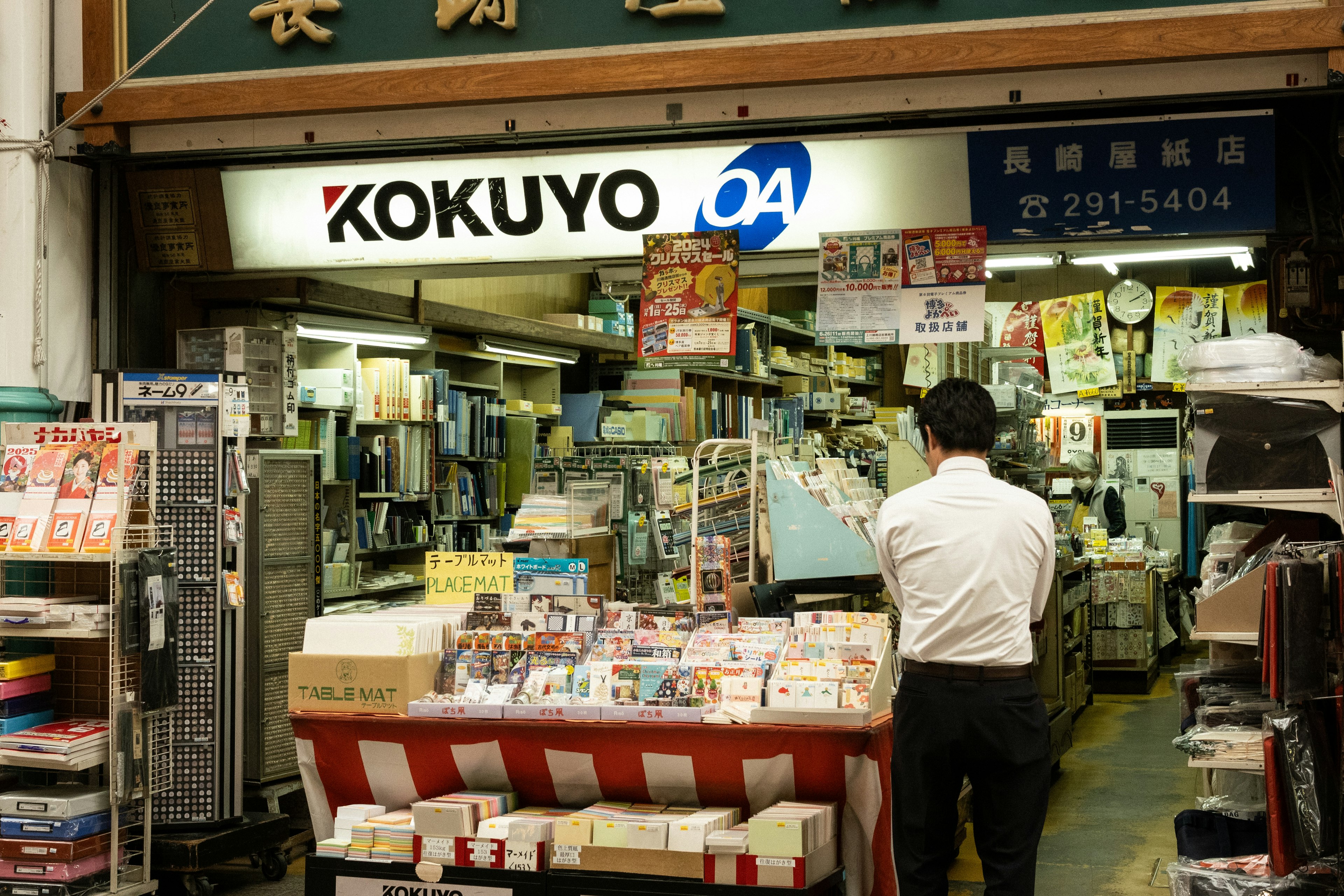 Fachada de KOKUYO con un hombre revisando artículos de papelería y revistas