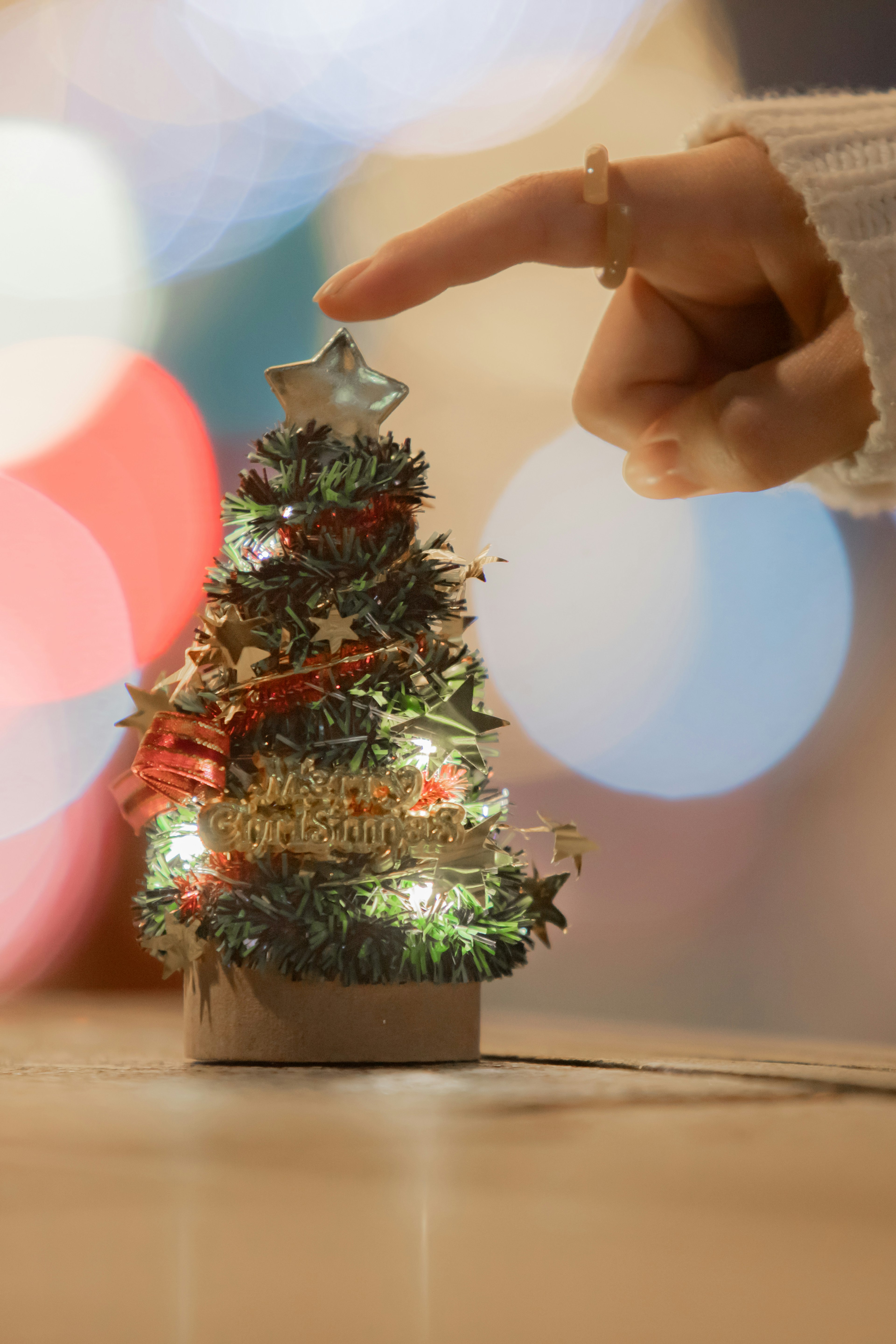 A hand reaching to touch a small Christmas tree with colorful bokeh lights in the background