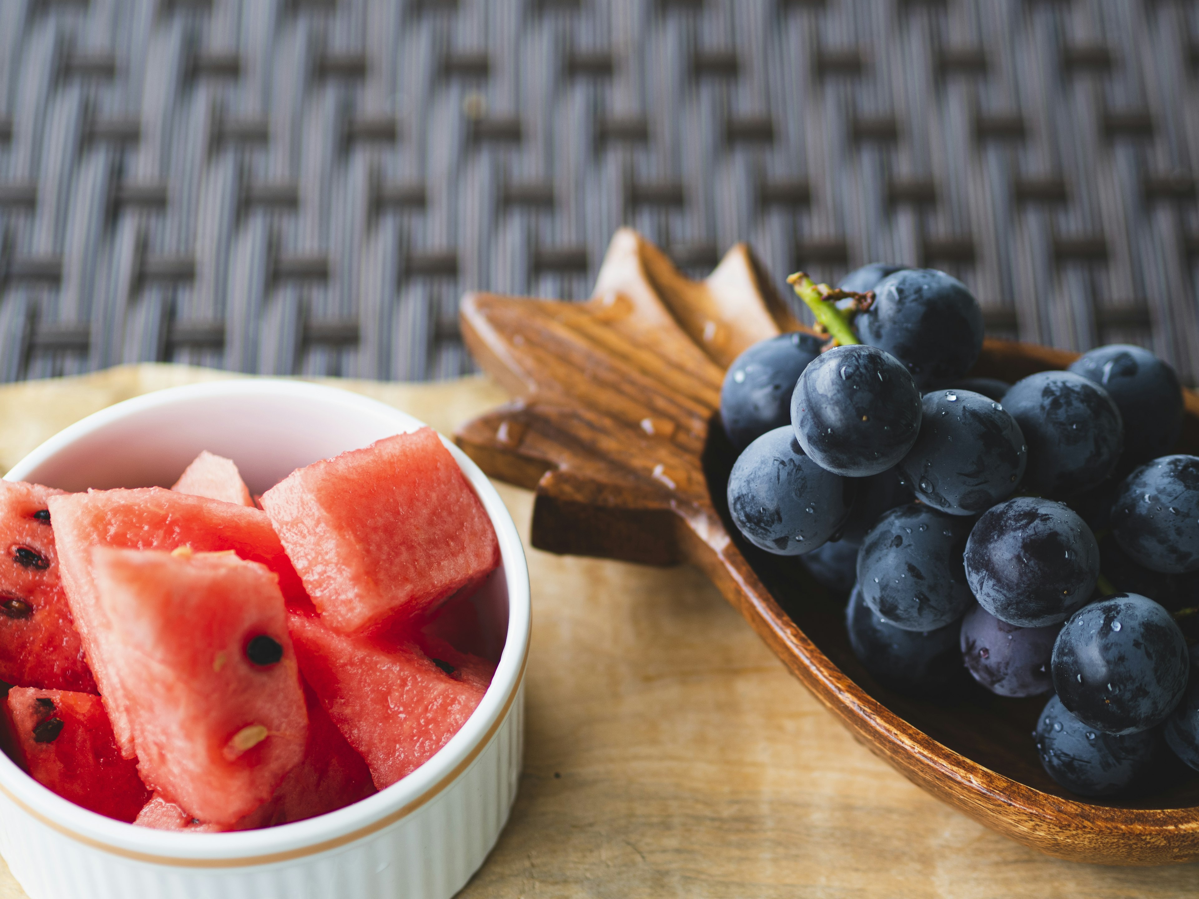 A white bowl with diced red watermelon and black grapes in a wooden dish