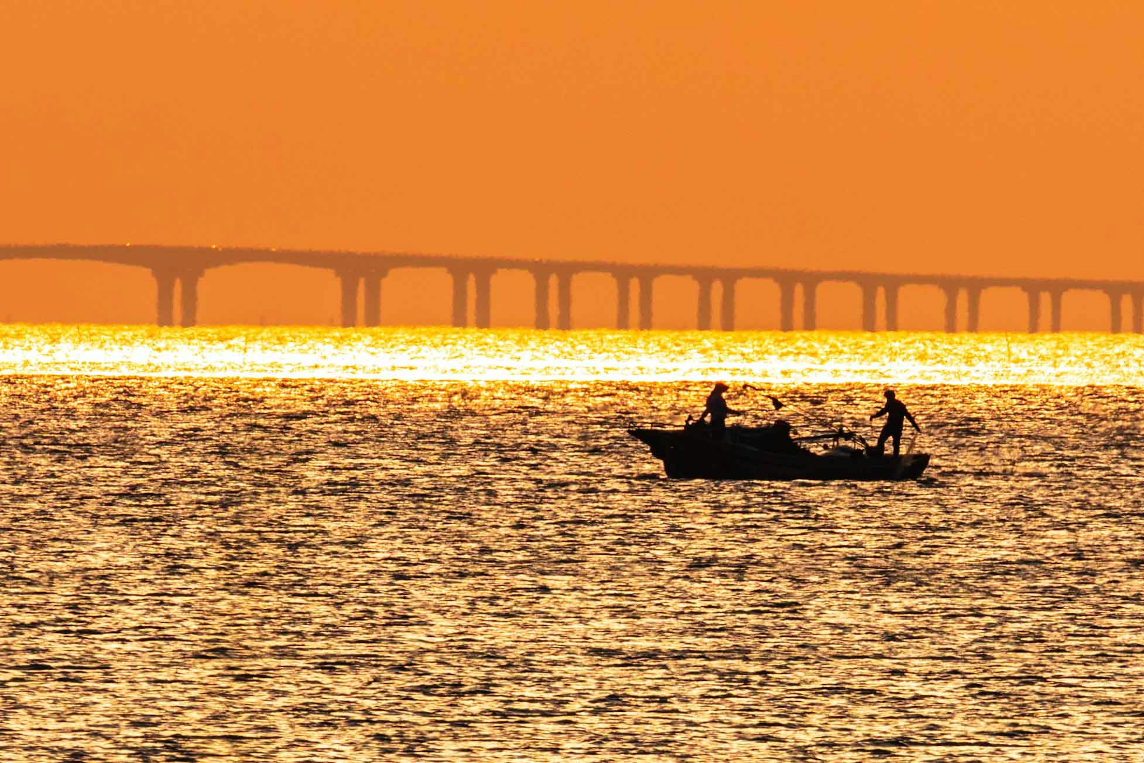 Silhouette di due pescatori su una barca contro un tramonto arancione con un ponte sullo sfondo