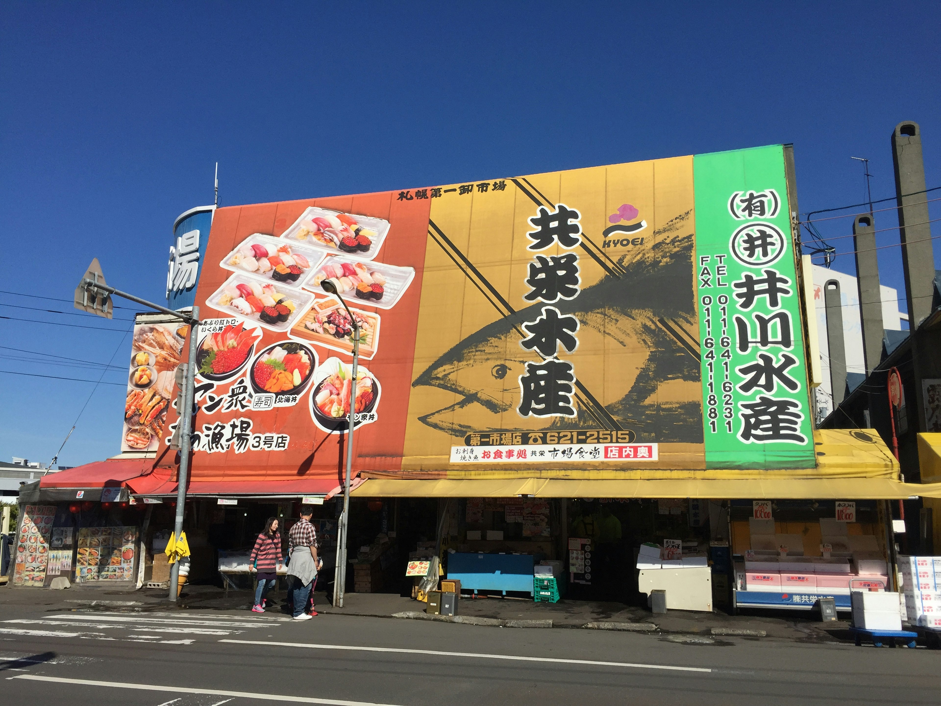 ภายนอกของร้านซูชิที่มีป้ายสีสันสดใสคึกคักใต้ท้องฟ้าสีฟ้า