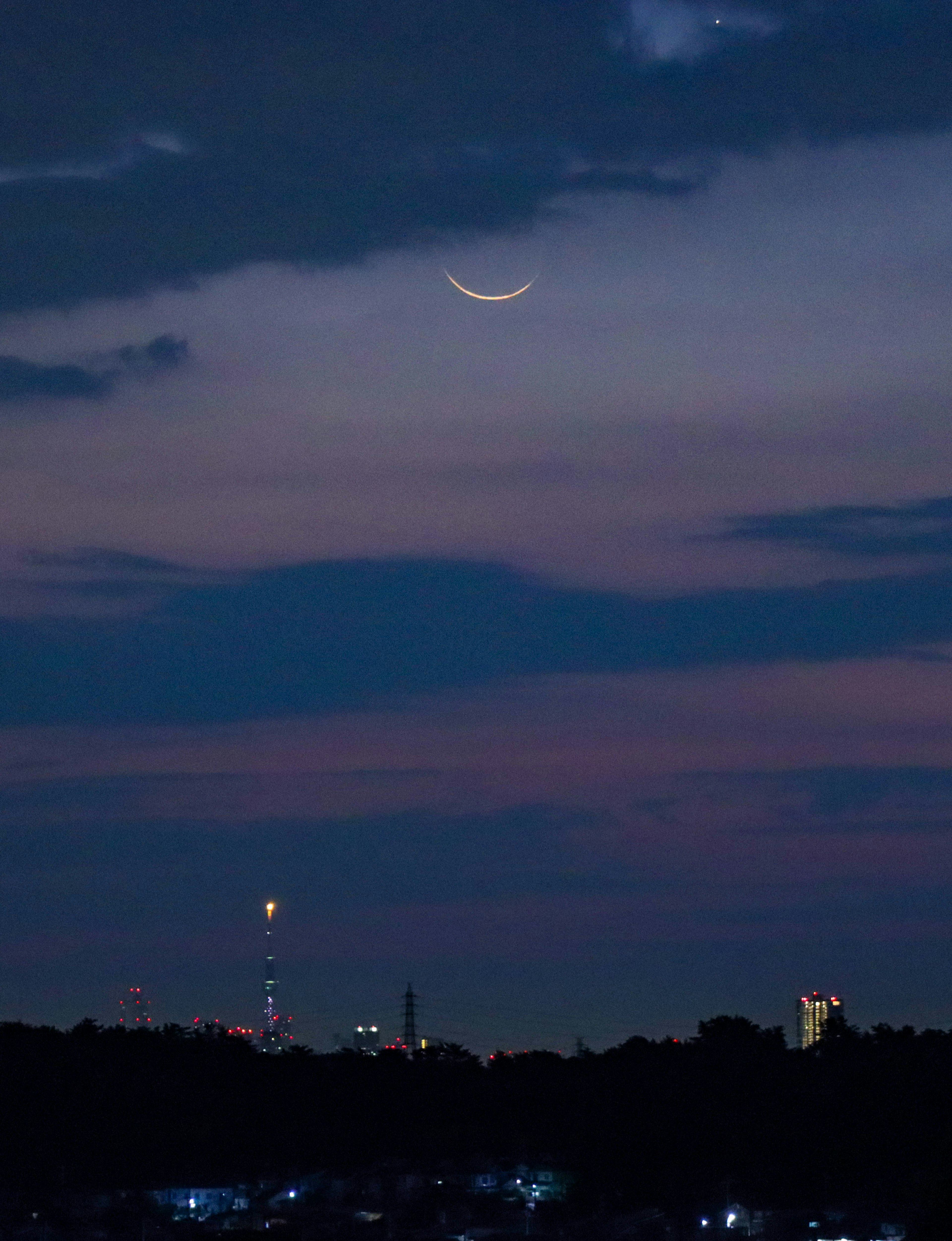 A crescent moon in a twilight sky with city silhouettes in the background