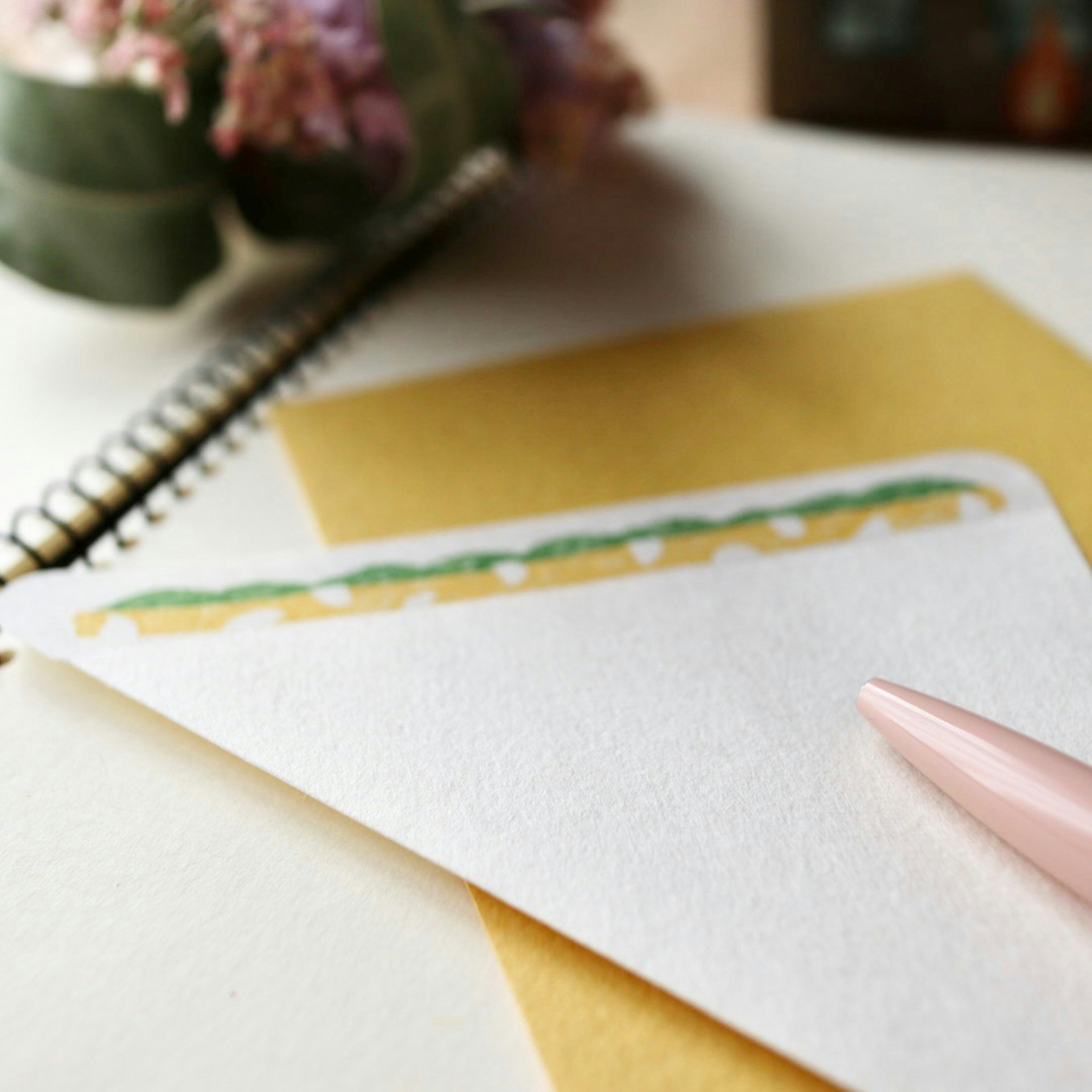 A desk scene featuring a white envelope with green design, a pink pen, and a yellow notepad