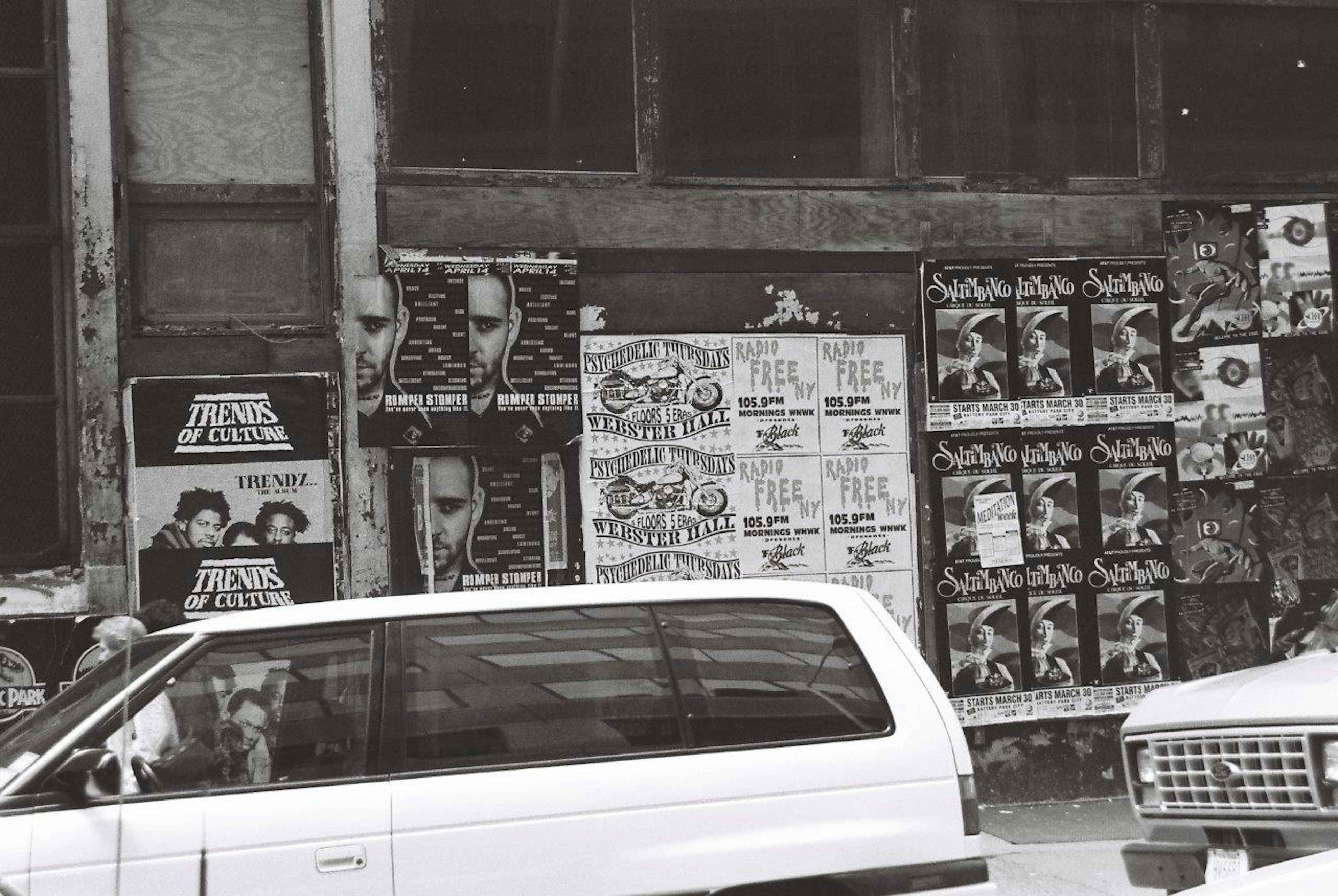 Scène de rue en noir et blanc avec des affiches de films sur un mur et une voiture blanche en circulation