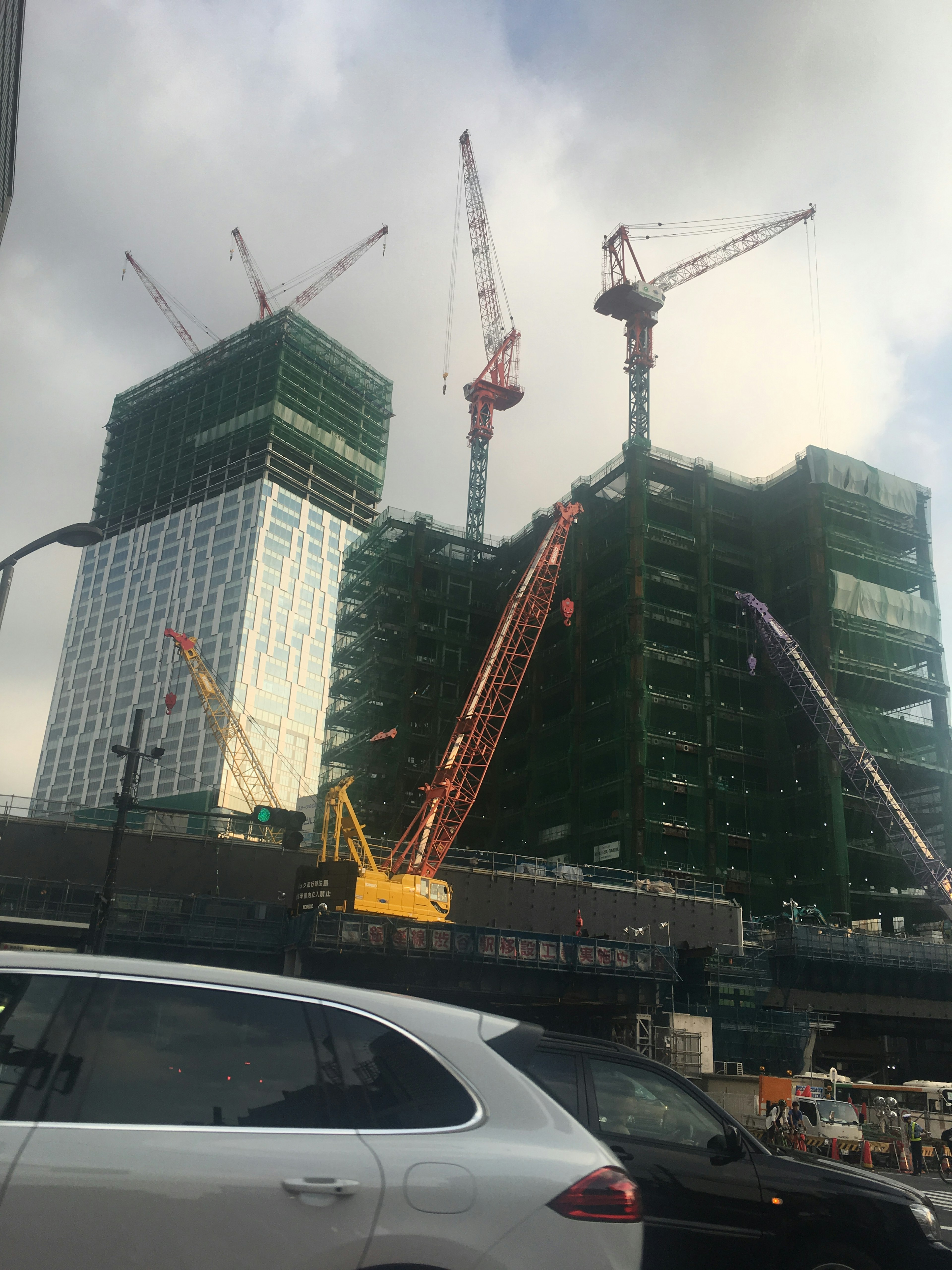 Construction site featuring tall buildings and cranes in the foreground a car is visible with green netting on the structures