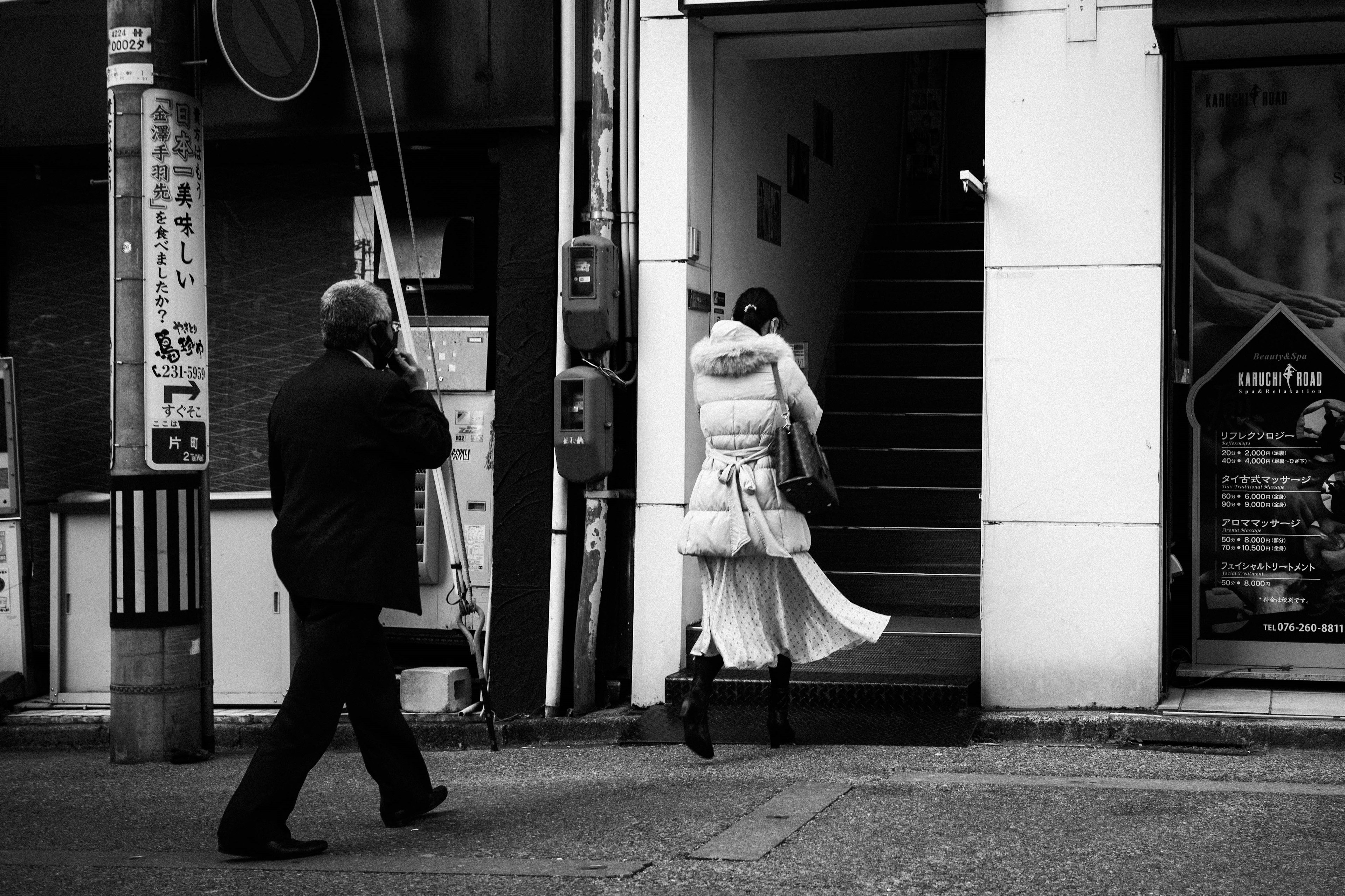 A woman in a white coat walking up stairs while a man in black is on the phone in a street scene