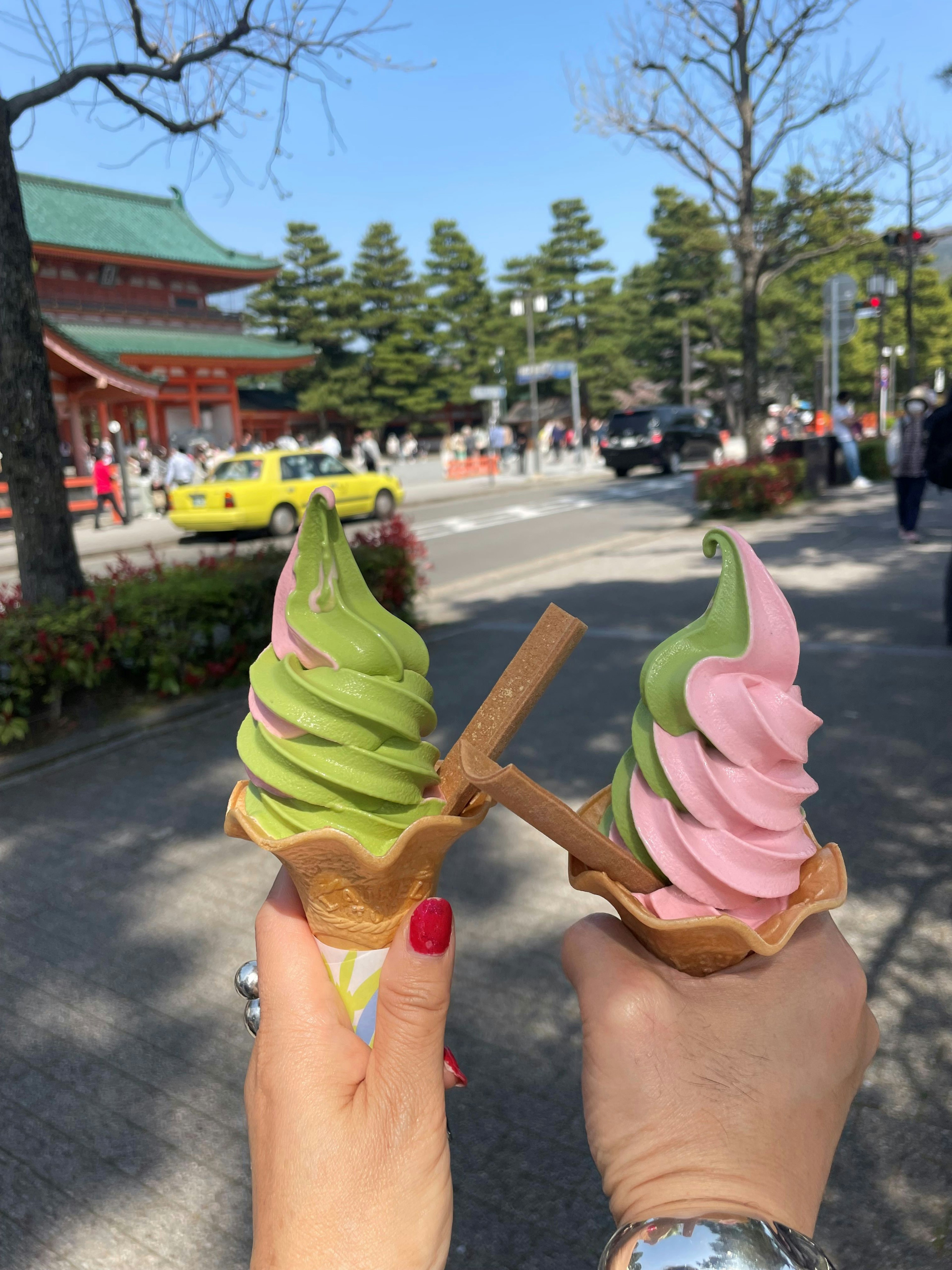 Dua tangan memegang kerucut es krim hijau dan merah muda dengan bangunan tradisional Jepang di latar belakang