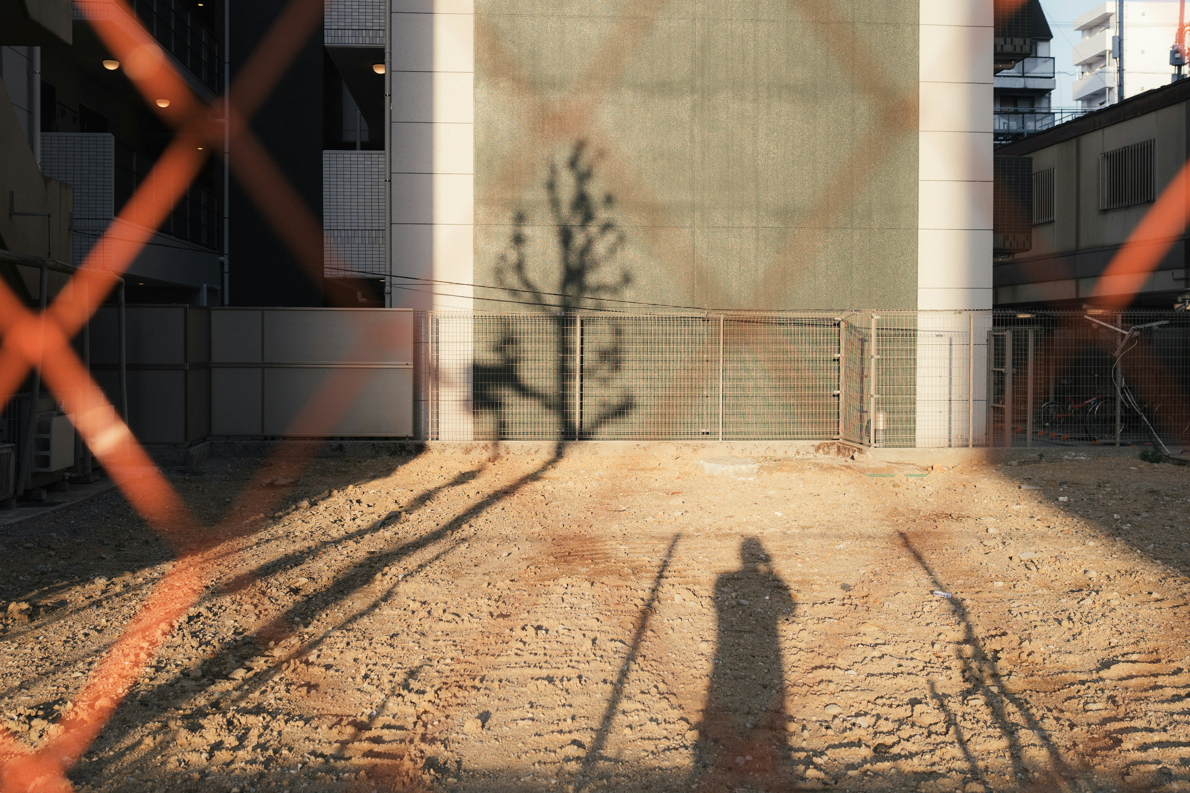 Ein Schotterplatz mit dem Schatten eines Baumes und dem Schatten einer Person hinter einem Zaun