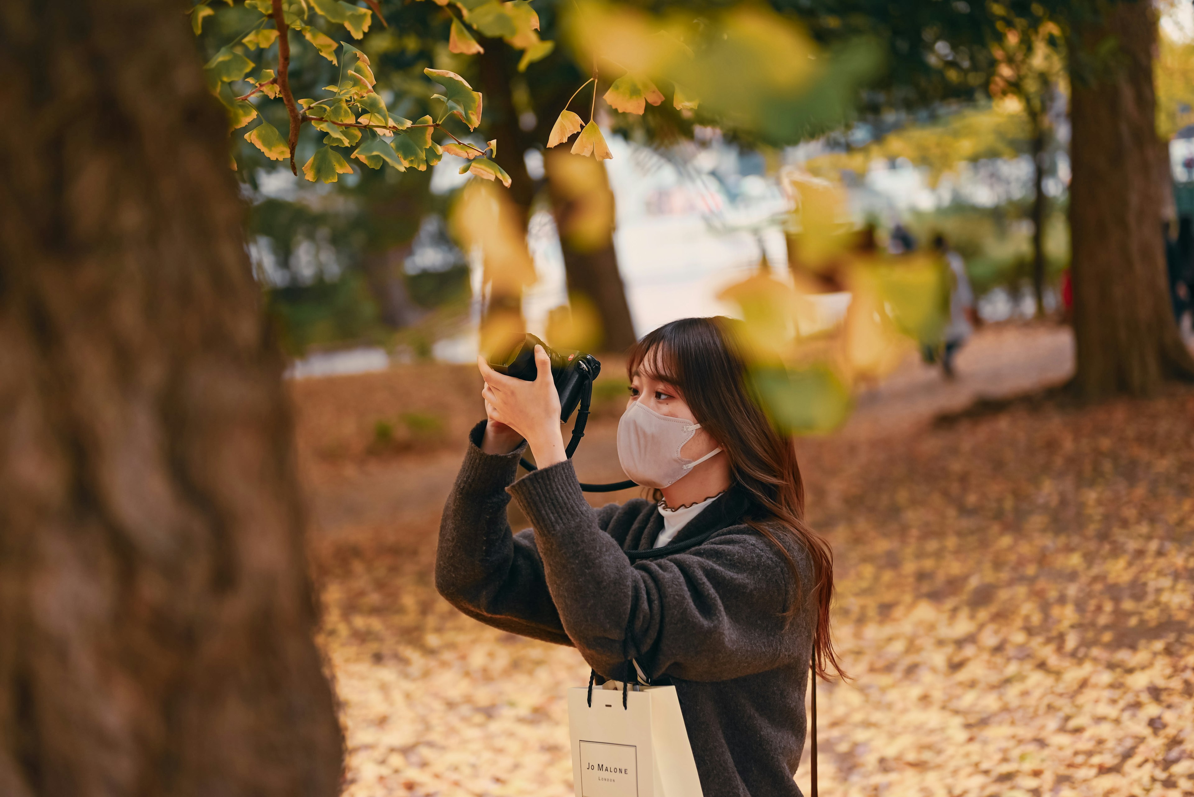 Femme tenant un appareil photo dans un parc d'automne portant un masque entourée d'arbres et de feuilles tombées