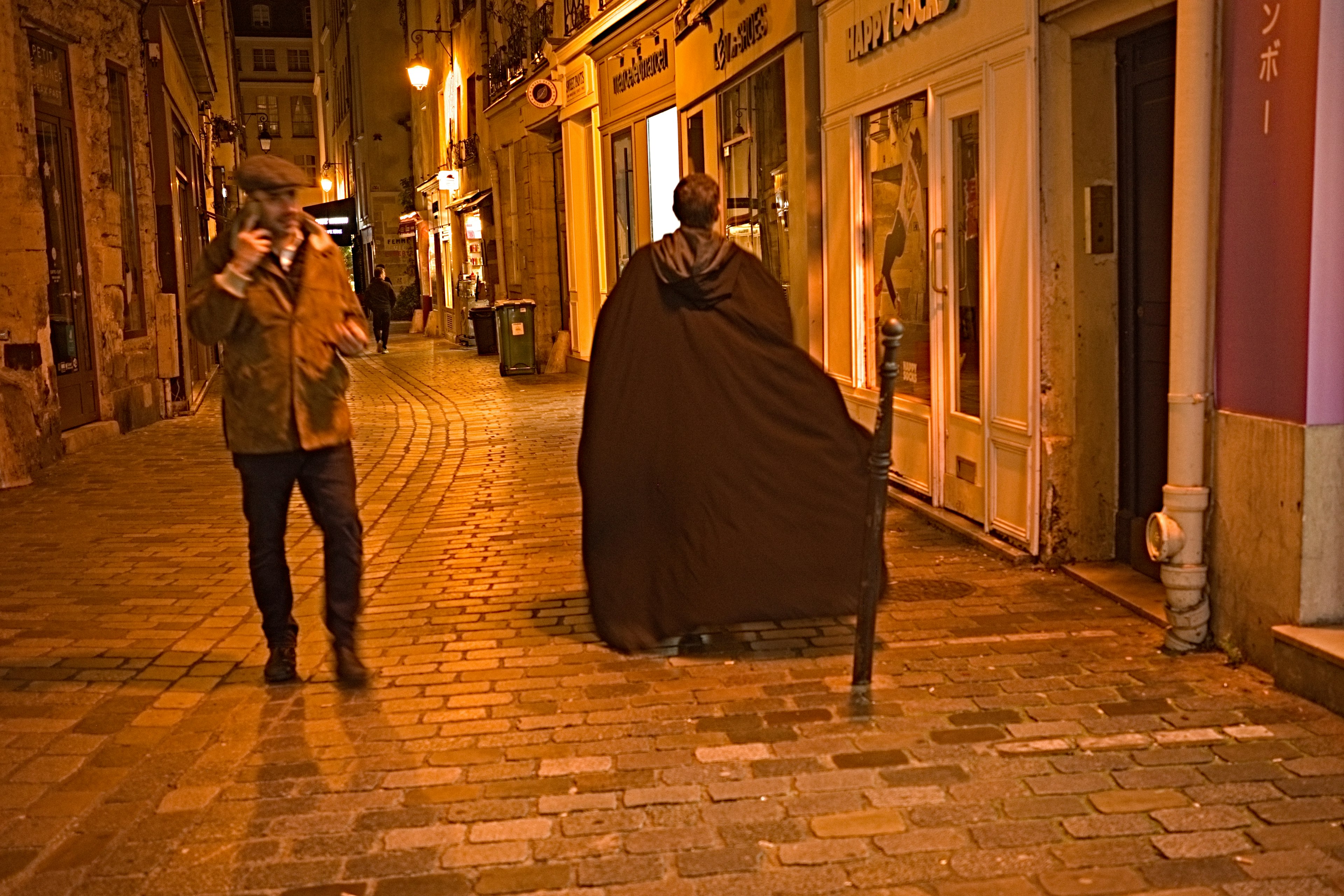 Person in a cloak walking in a nighttime street with another person holding a camera