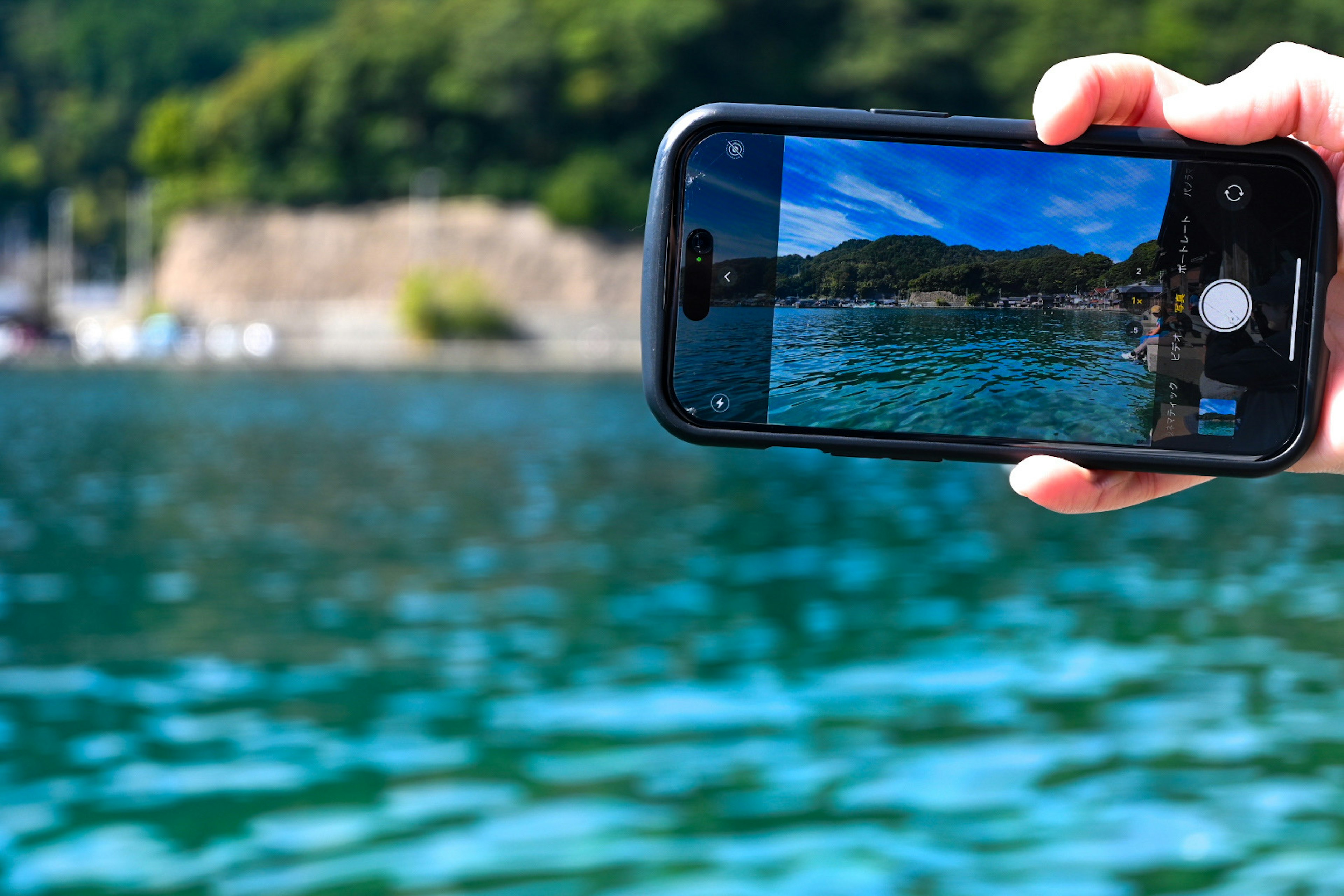 Eine Person hält ein Smartphone und fotografiert eine Landschaft mit Wasser im Vordergrund