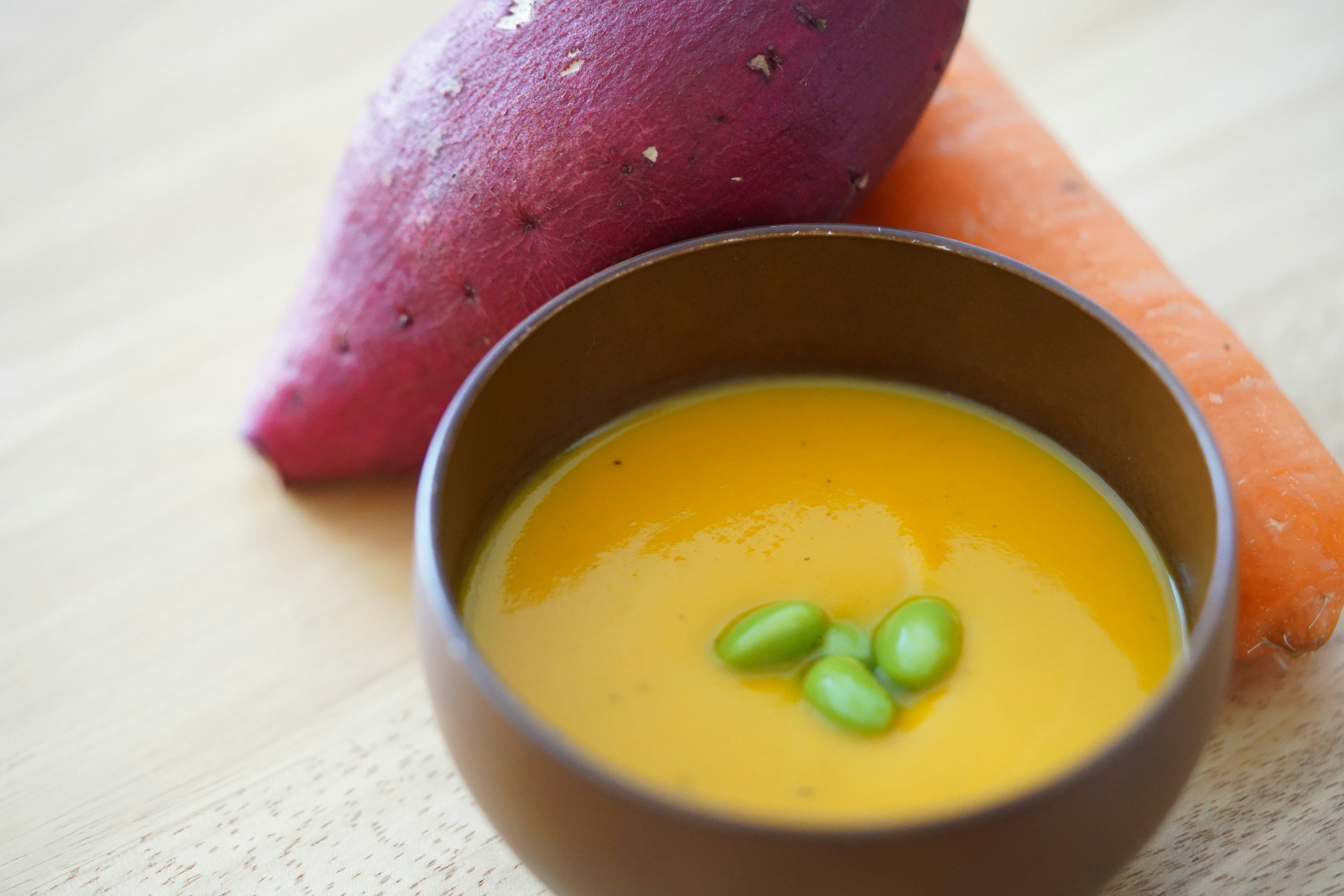 Schüssel mit orangefarbener Suppe garniert mit grünen Bohnen neben lila Süßkartoffel und Karotte