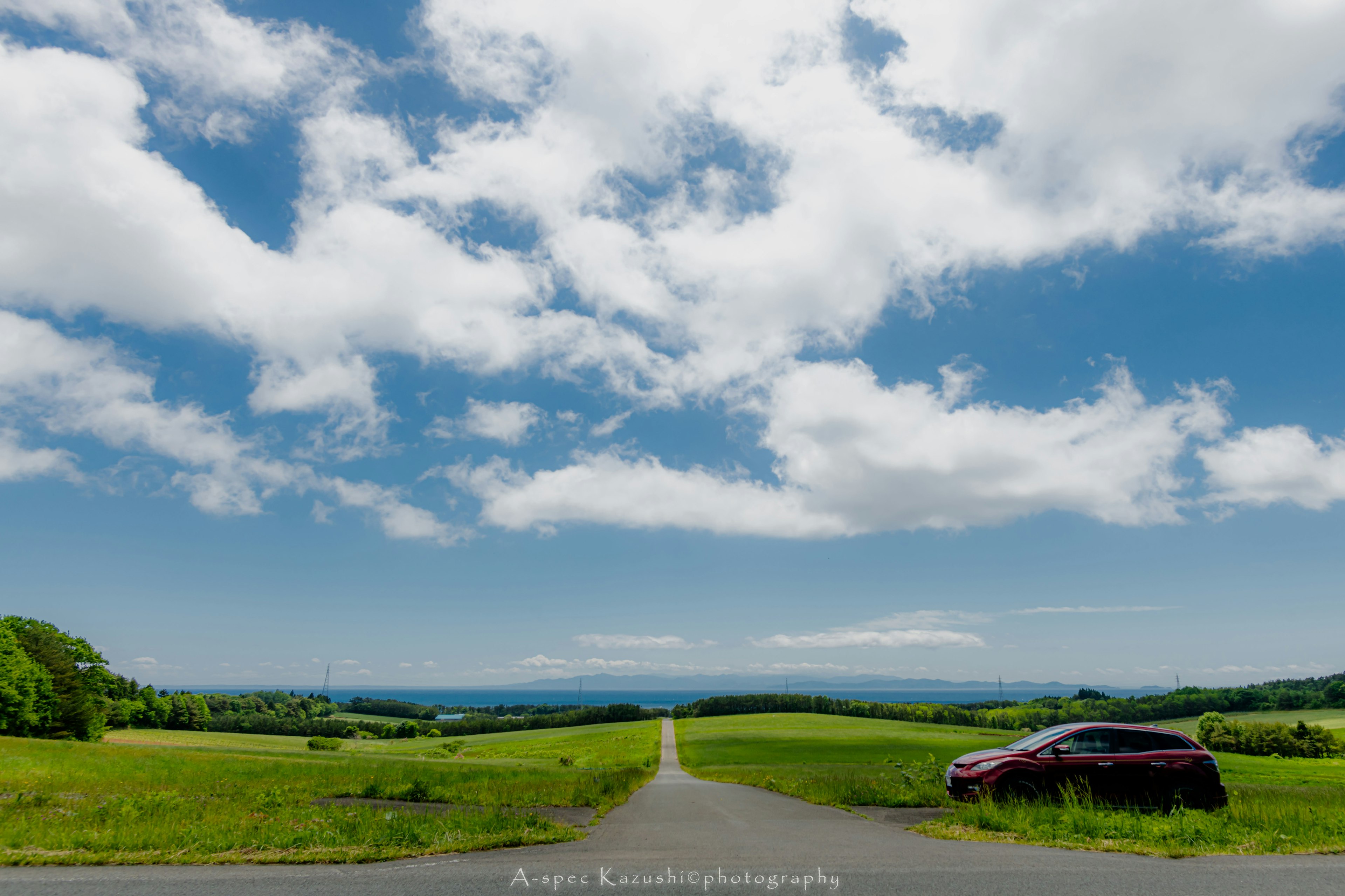 青空と雲が広がる緑の風景に沿った道路と赤い車