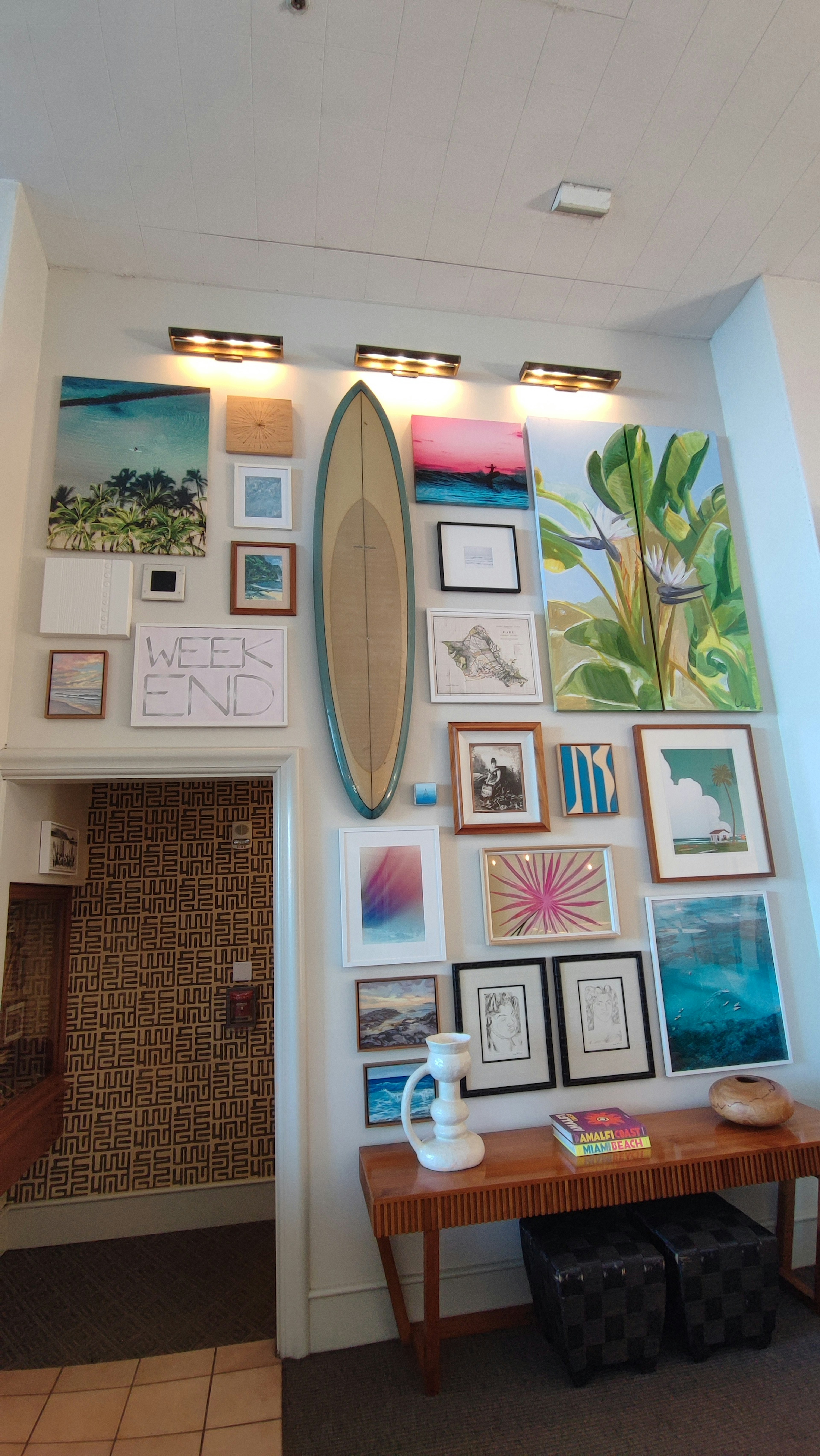 Wall display featuring a surfboard and various framed artworks with a wooden table