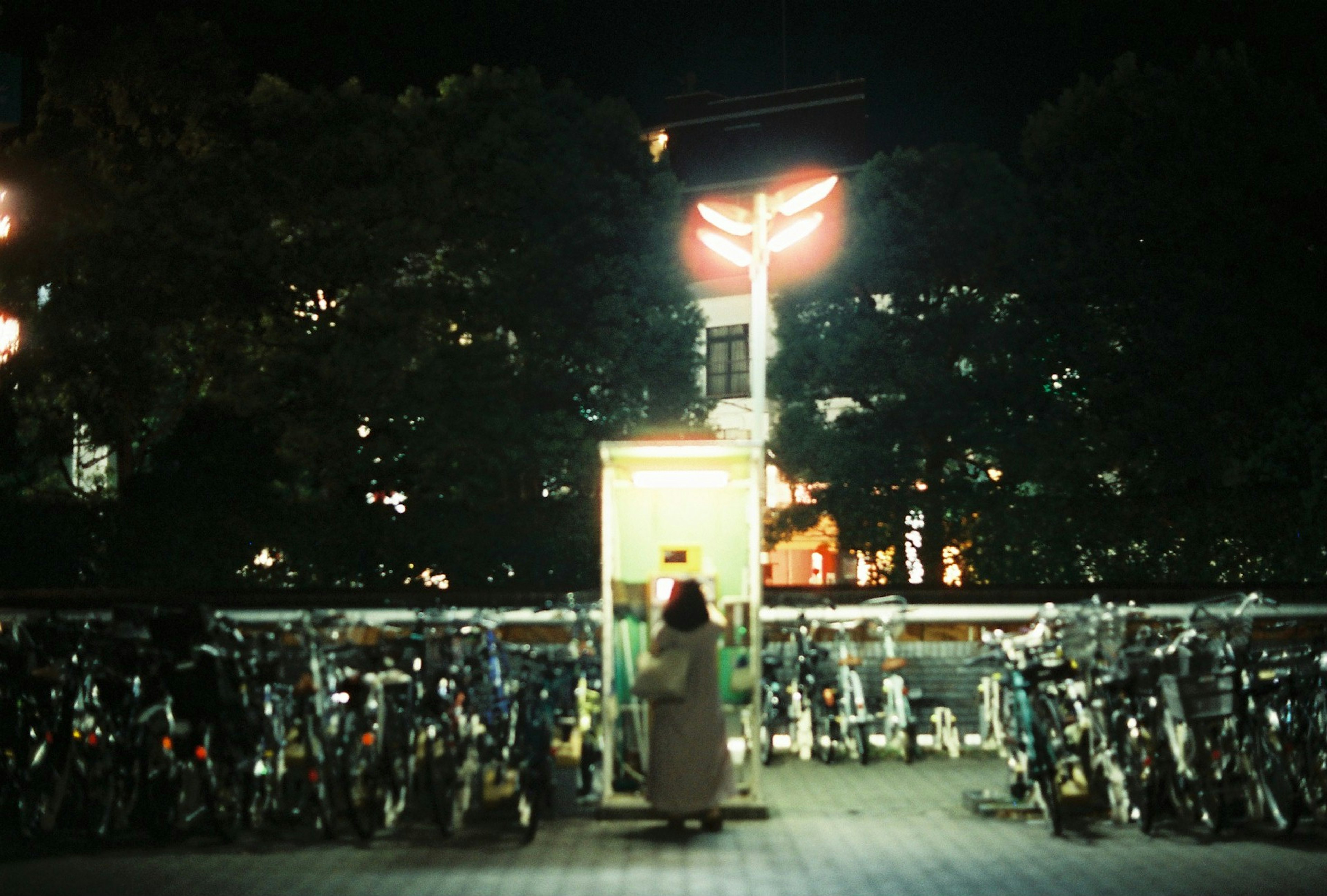 Una figura de pie frente a una cabina telefónica por la noche rodeada de bicicletas