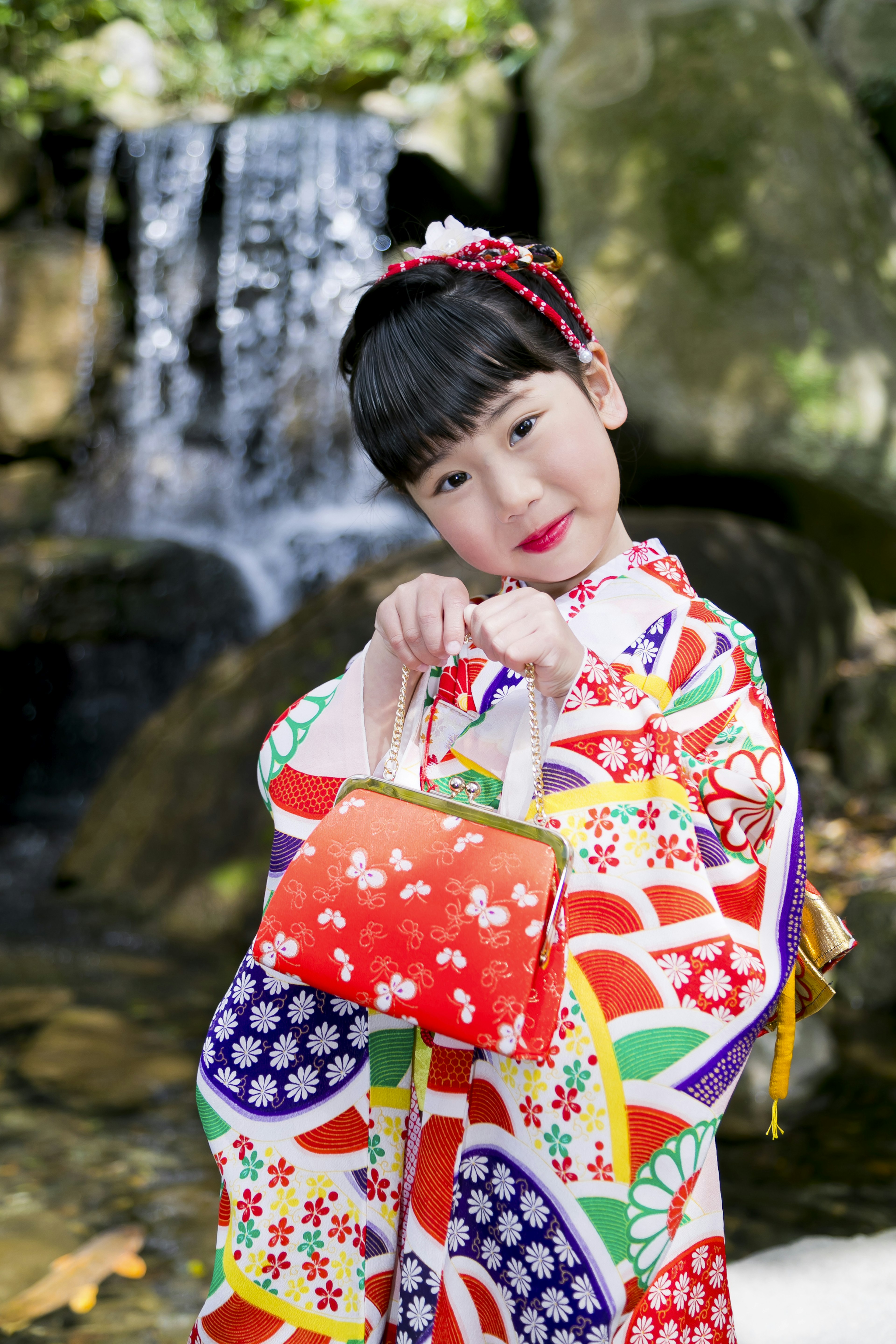 Una niña en kimono colorido sosteniendo una pequeña bolsa roja cerca de una cascada