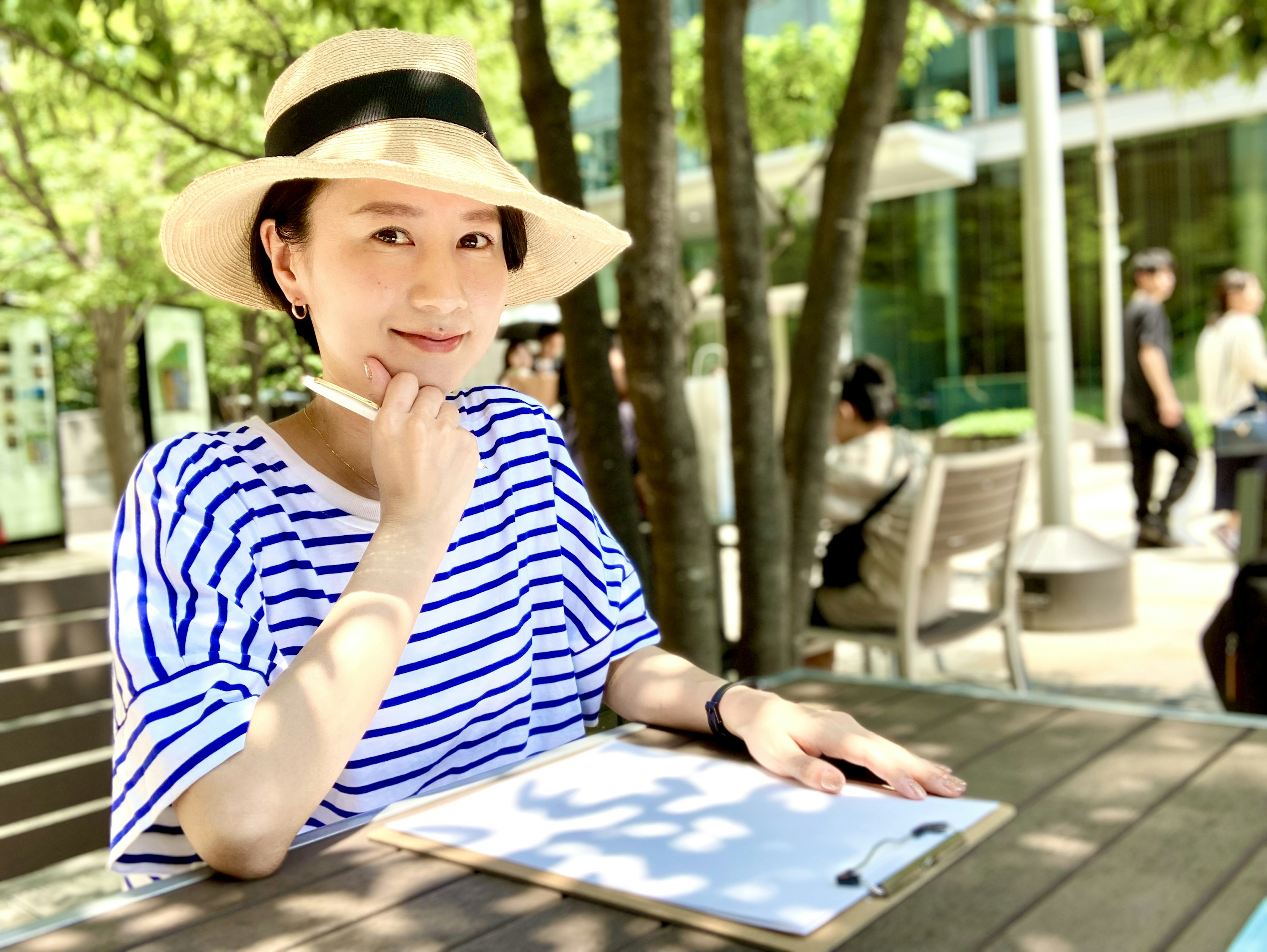 A woman wearing a striped shirt and hat smiling at an outdoor table with a notebook