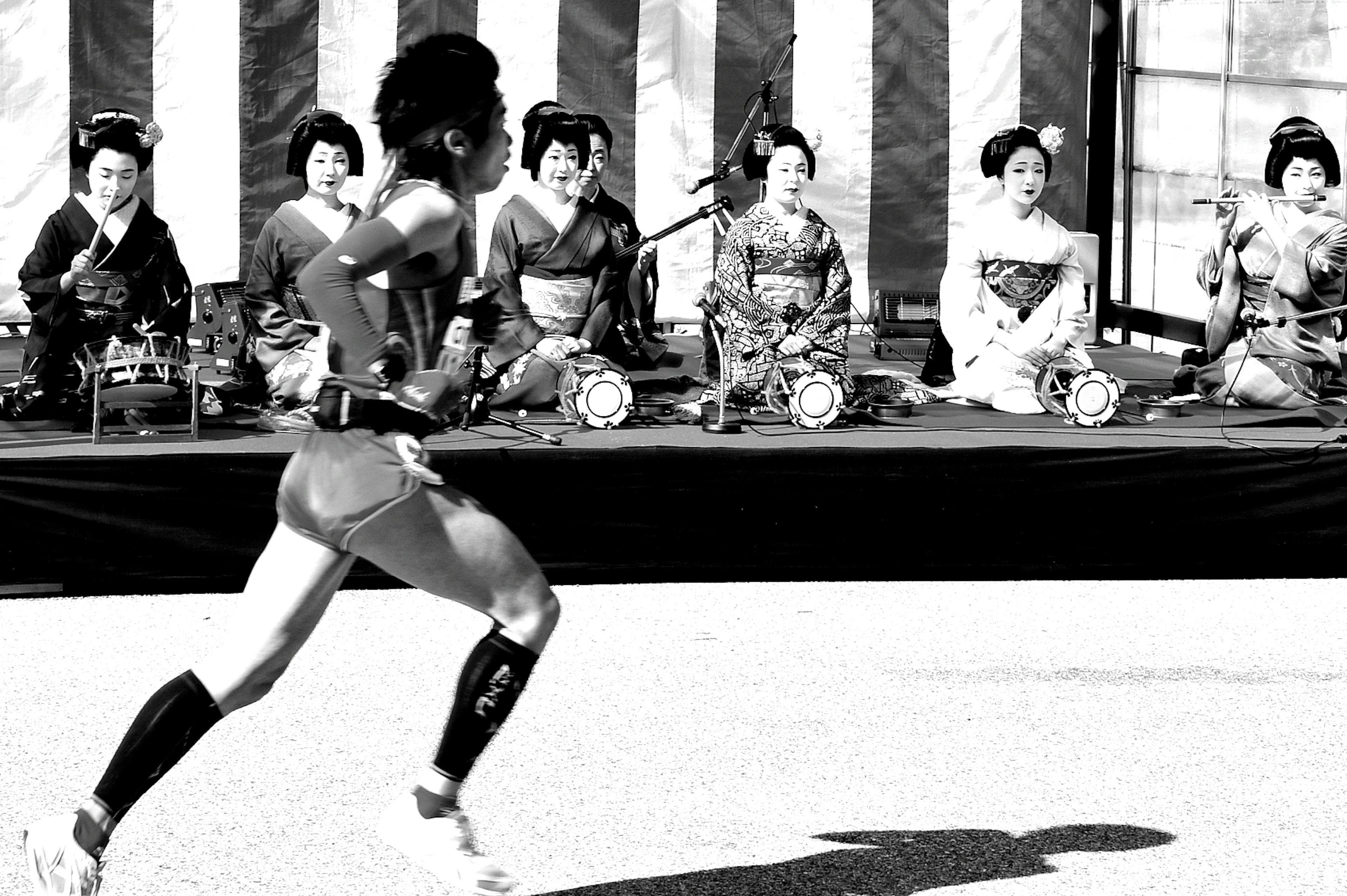 Un coureur en mouvement avec des femmes en tenue traditionnelle assises en arrière-plan dans une image en noir et blanc