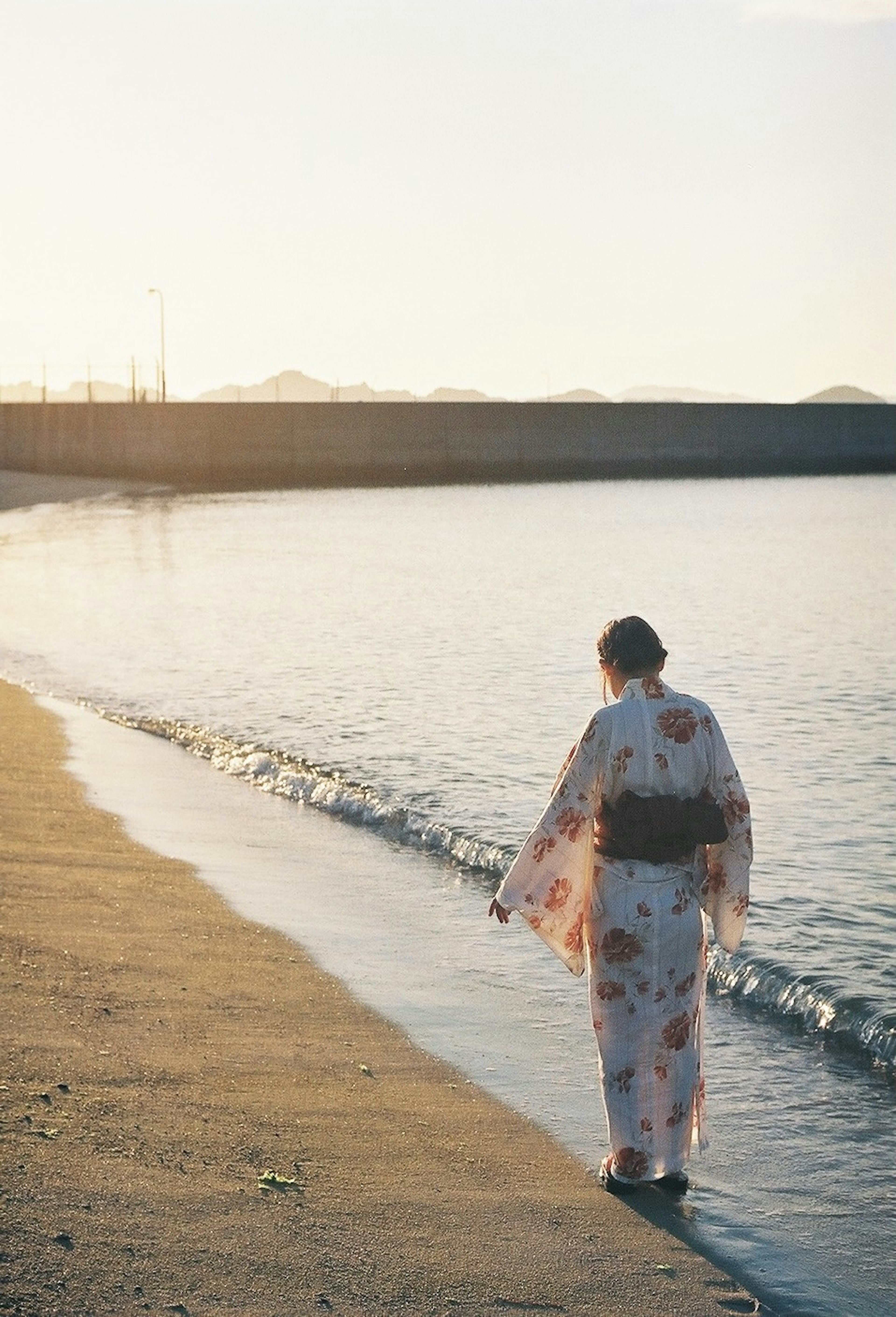 夕日を背にした着物姿の女性が海岸を歩いている