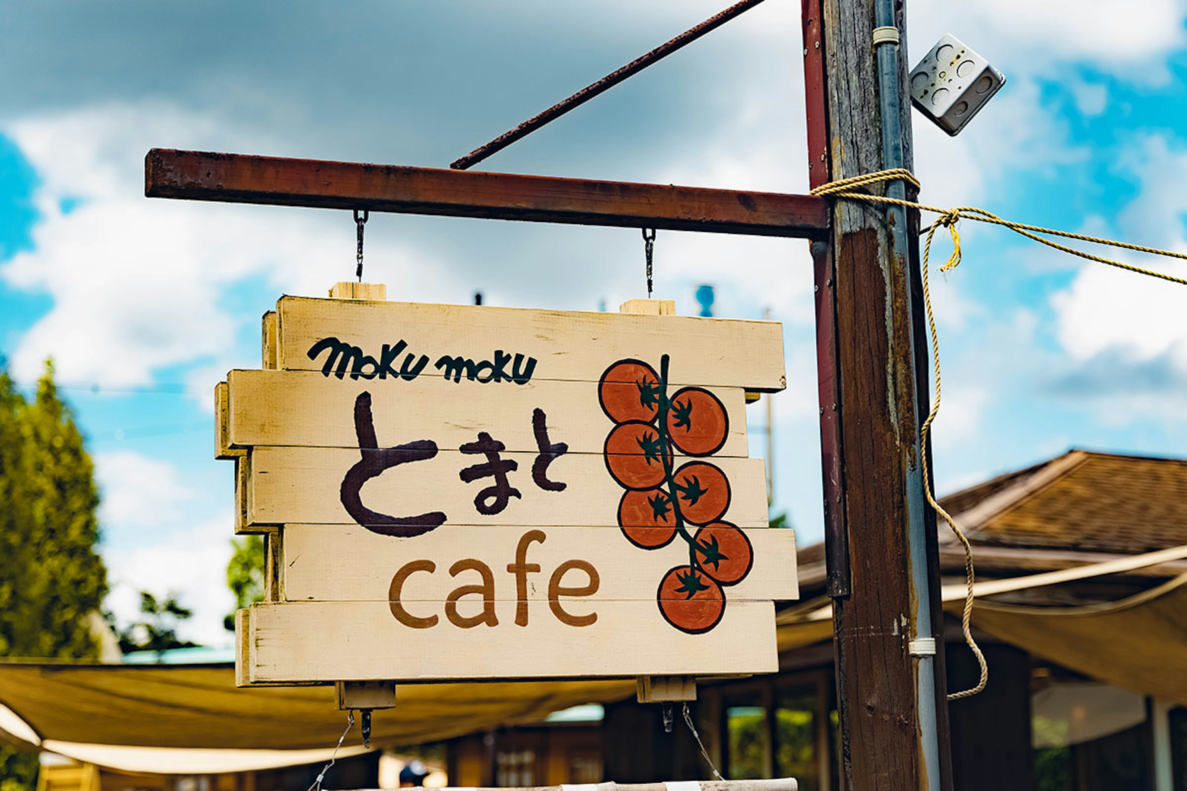 Cartel de café con las palabras 'moku moku' y 'とまと' con un fondo de cielo azul y vegetación