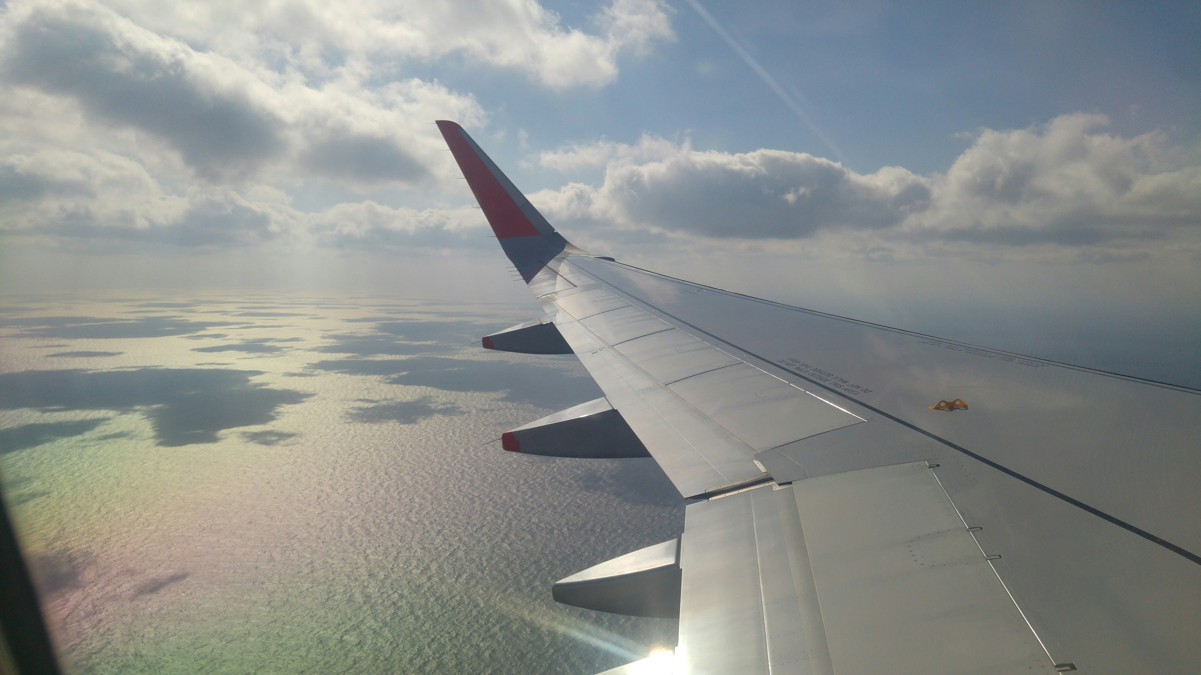 Bild zeigt einen Flugzeugflügel mit einer malerischen Aussicht auf das Meer farbige Reflexionen unter einem blauen Himmel mit Wolken