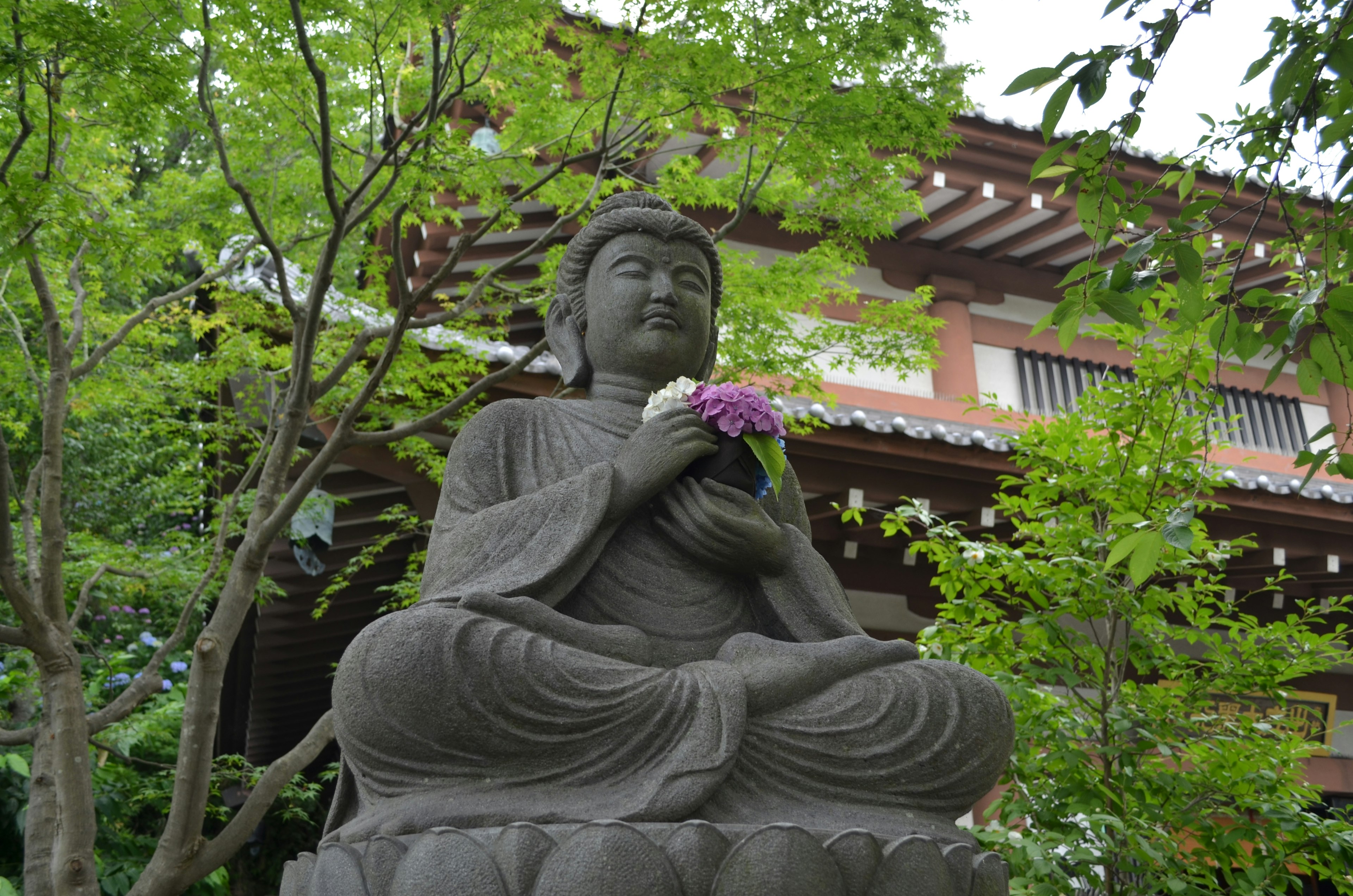 Statua del Buddha seduta tra alberi verdi con un fiore in mano e un'espressione serena