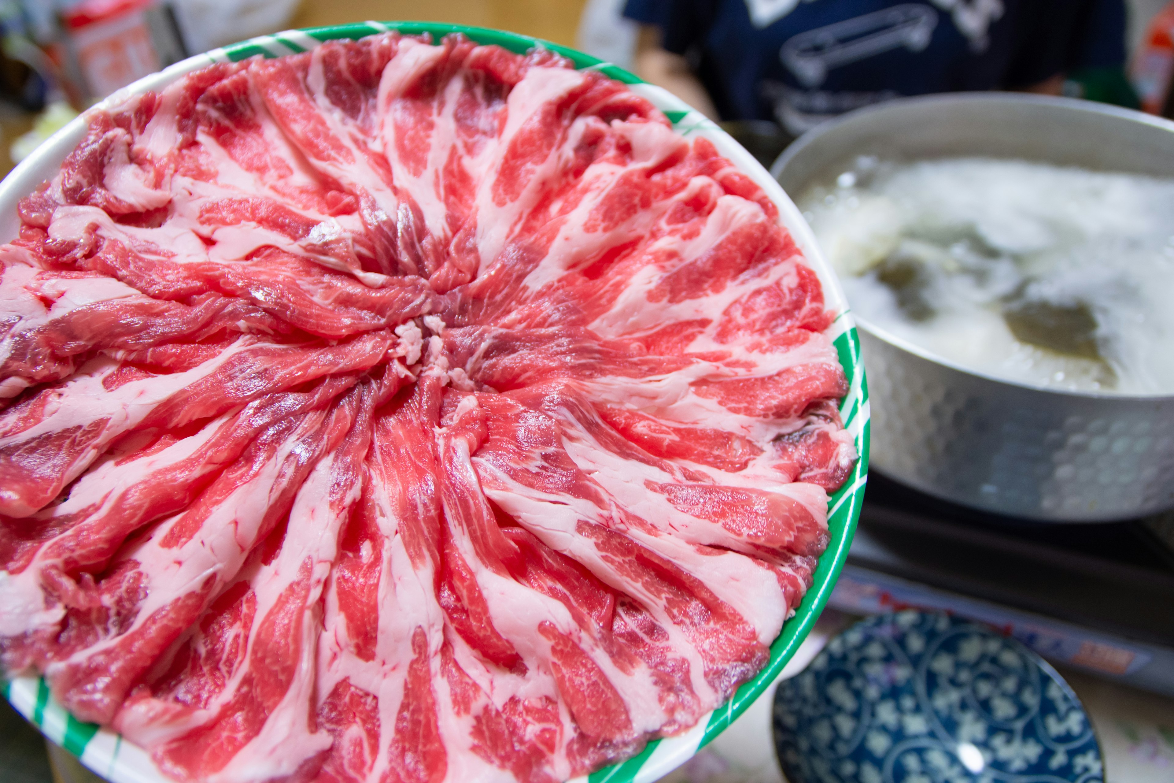 Un plato de carne en rodajas finas dispuestas en un patrón circular con una olla de agua hirviendo